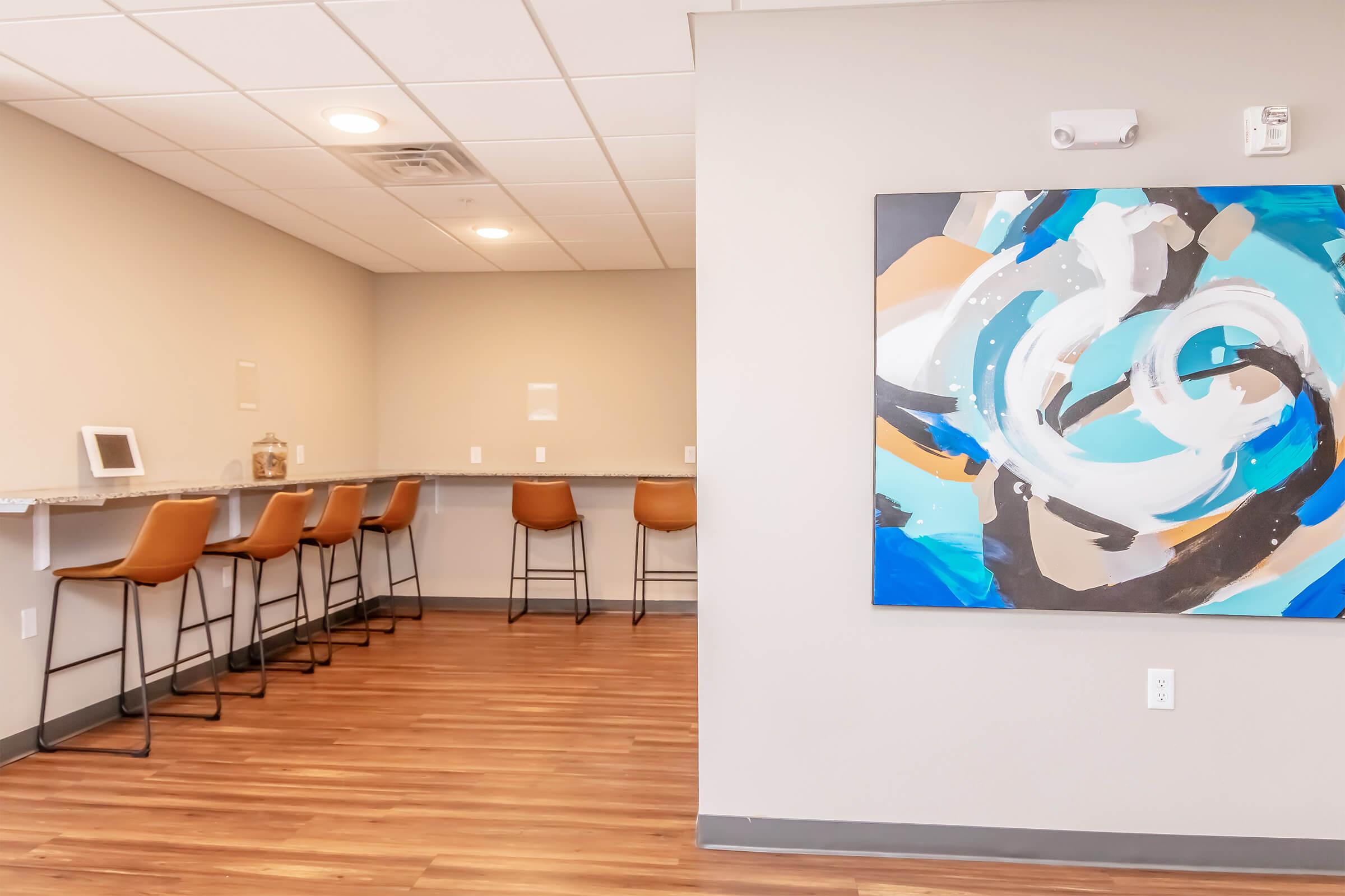 A modern, minimalistic interior featuring a bar-style seating area with high stools along a gray countertop. In the background, a large abstract painting with blue, white, and orange swirls adds a pop of color against the neutral walls and wooden flooring.