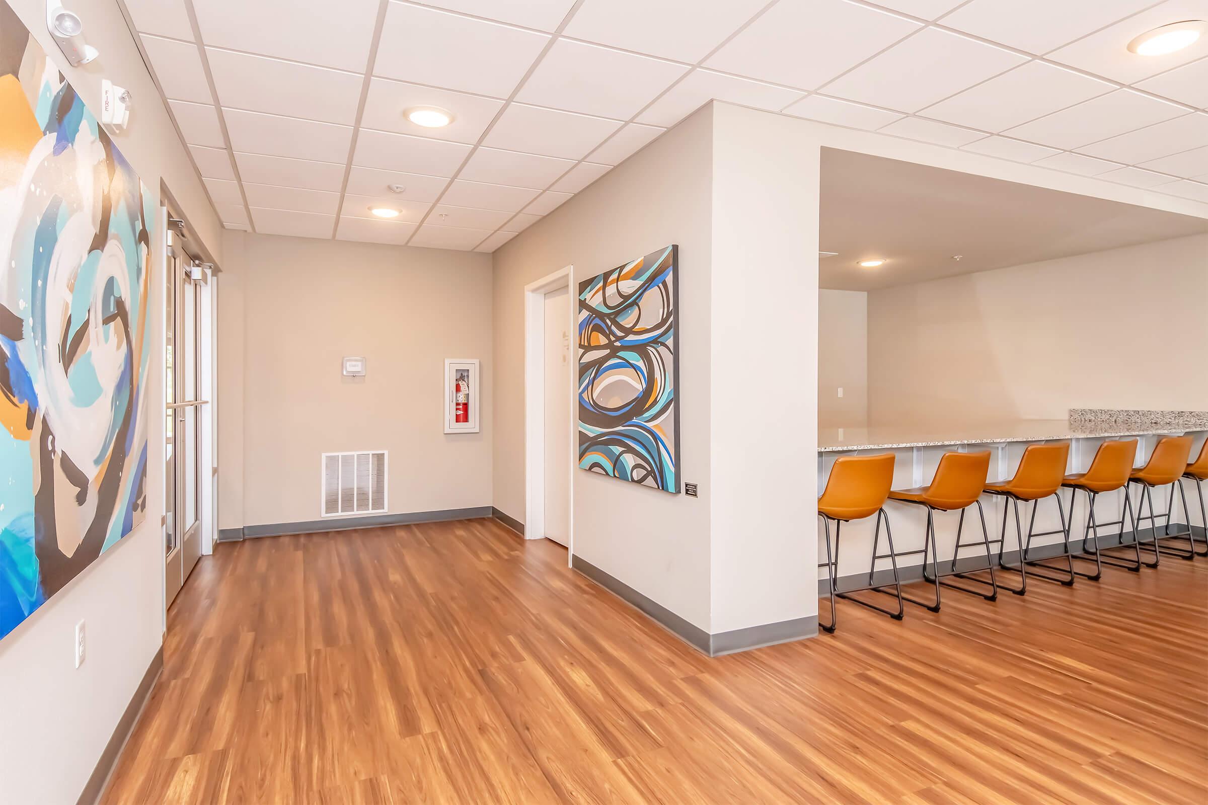 A modern interior space featuring a wooden floor, light-colored walls, and a ceiling with grid tiles. On one side, there are colorful abstract paintings on the wall, while the other side includes a long countertop with bar stools. The space is well-lit with natural light from large windows.