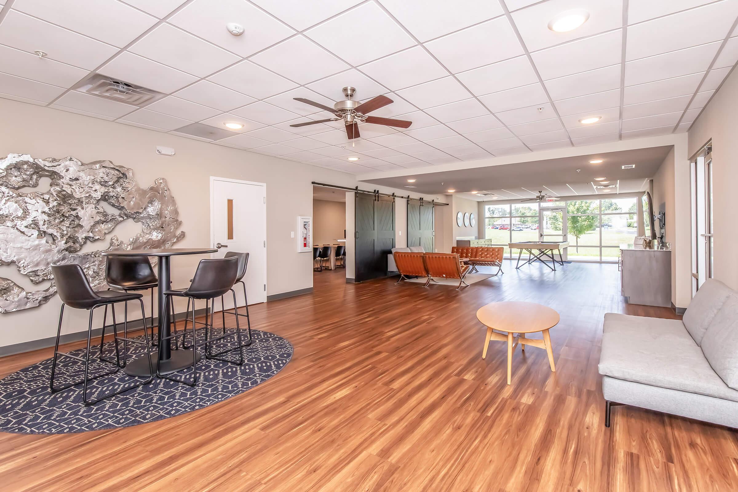 A spacious, modern living area featuring a ceiling fan, hardwood flooring, and minimalist decor. There is a round table with four high chairs, a cozy sofa, an orange loveseat, and a small round coffee table. Glass panels separate the room from an outdoor view, creating an open and inviting atmosphere.