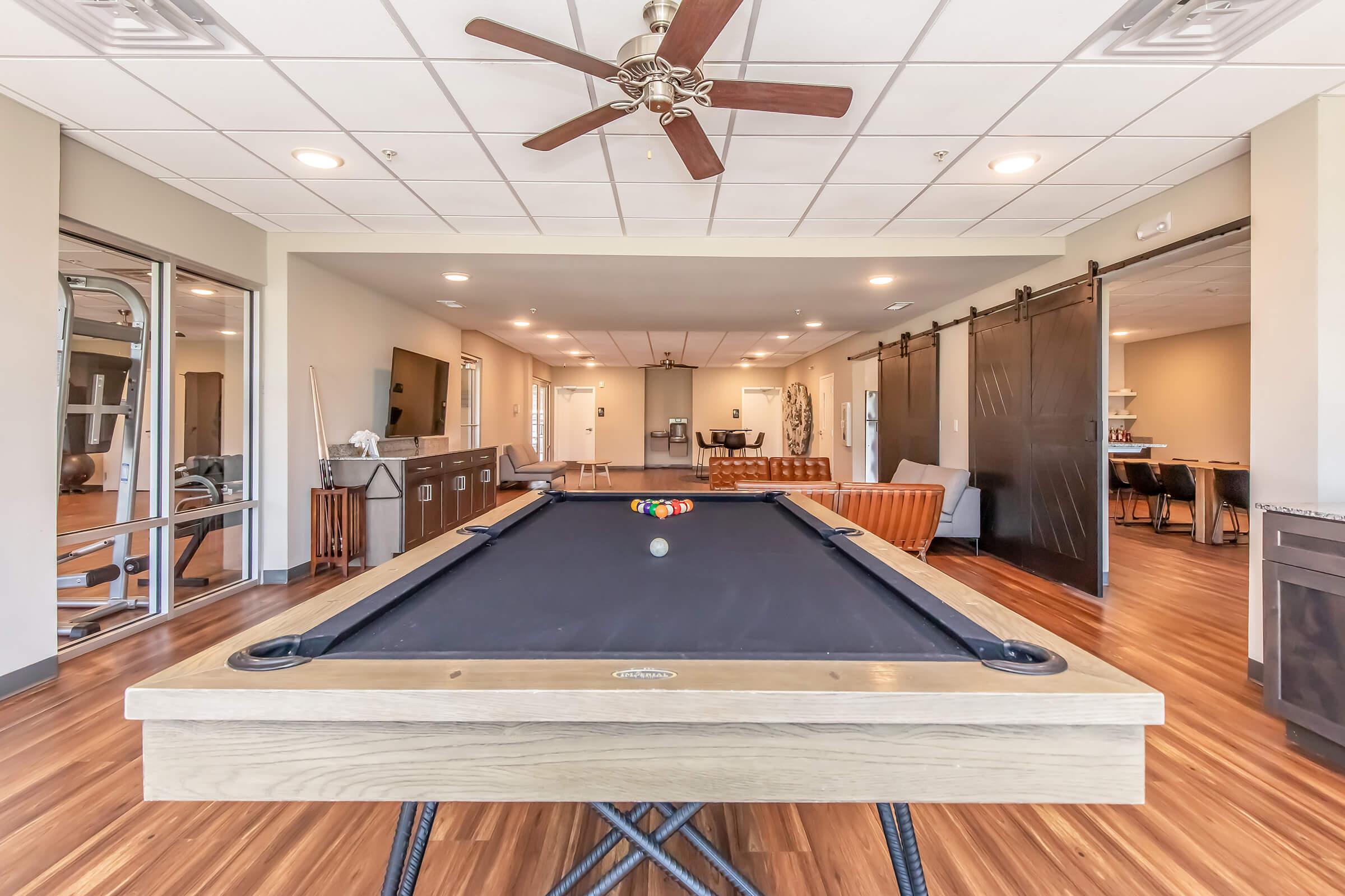 A spacious recreational room featuring a pool table in the foreground, with a ceiling fan overhead. The background shows a modern lounge area with a sofa and a large window. The room is well-lit with wooden flooring, creating a comfortable atmosphere for leisure activities.