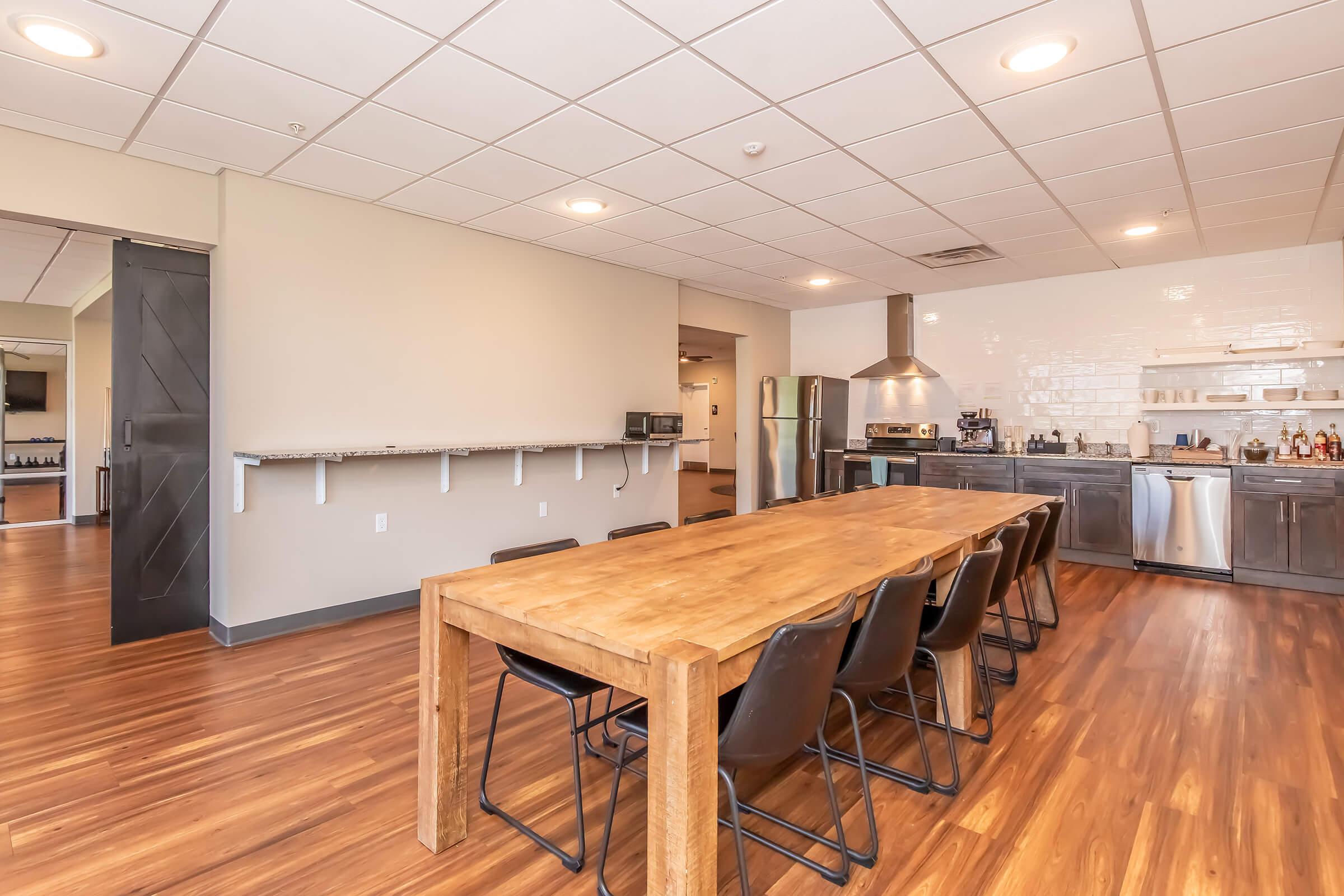 A spacious modern kitchen with a long wooden dining table and black chairs. The kitchen area features stainless steel appliances, including a refrigerator and a stove. The floor is made of hardwood, and the walls are painted light colors, creating a bright and inviting atmosphere.