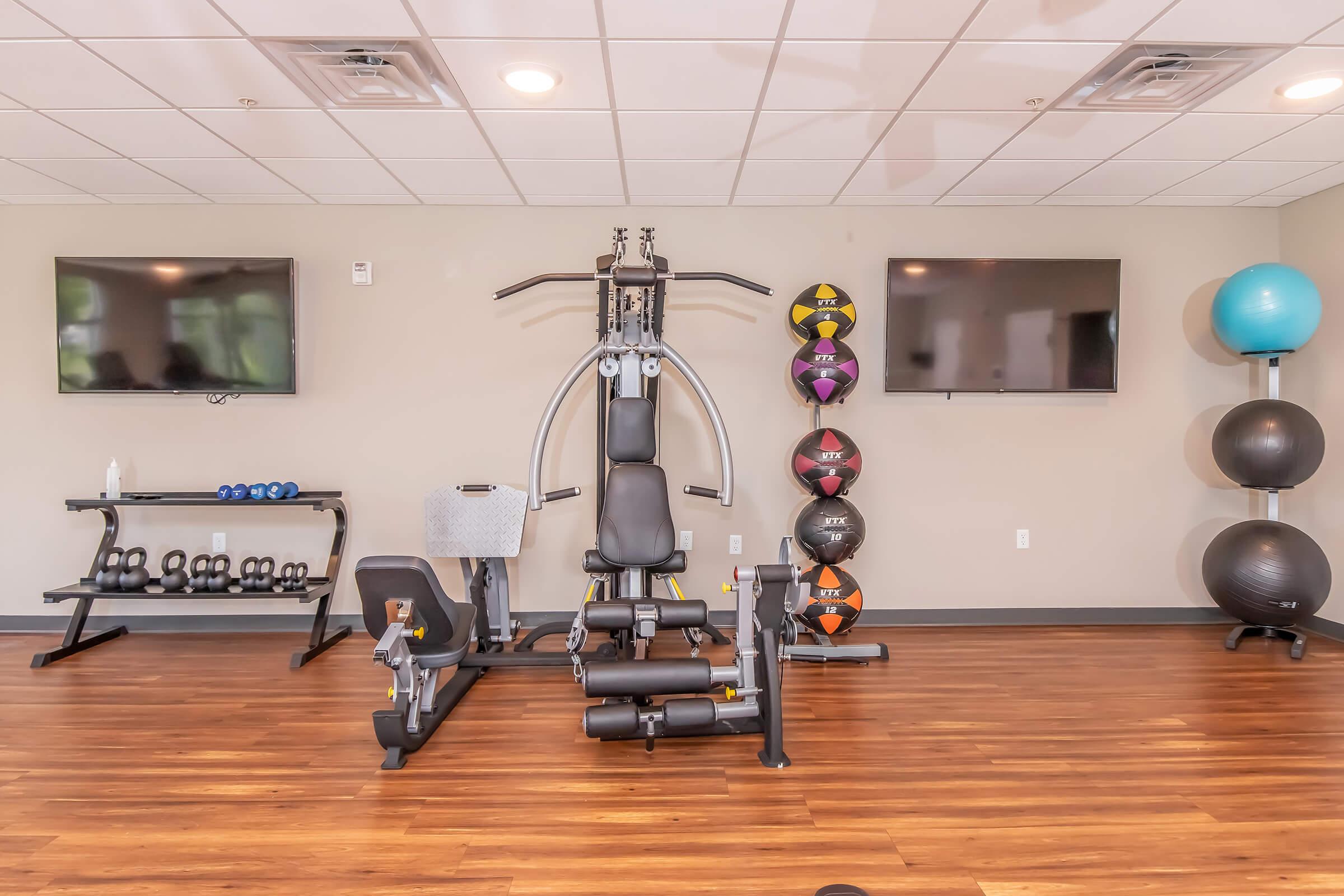 A modern fitness room featuring a multi-station gym machine, two flat-screen TVs on the wall, and a variety of exercise equipment including dumbbells, stability balls, and medicine balls neatly arranged on shelves. The flooring is wooden, creating a warm and inviting atmosphere for workouts.