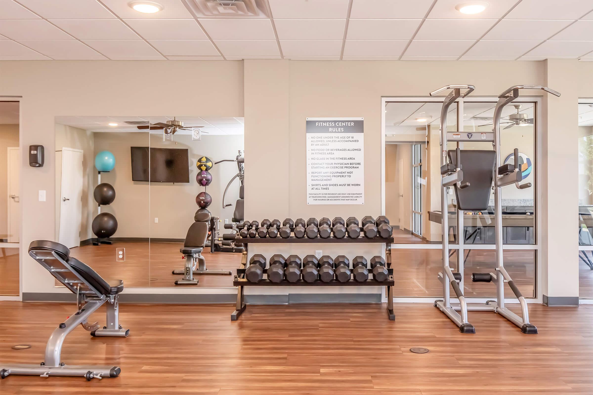 A modern fitness center with wooden flooring, featuring a range of dumbbells on a rack, a workout bench, and a multi-gym machine. Mirrors reflect the space, which includes exercise balls, a wall-mounted TV, and fitness guidelines displayed on the wall. The ambiance is bright and inviting.