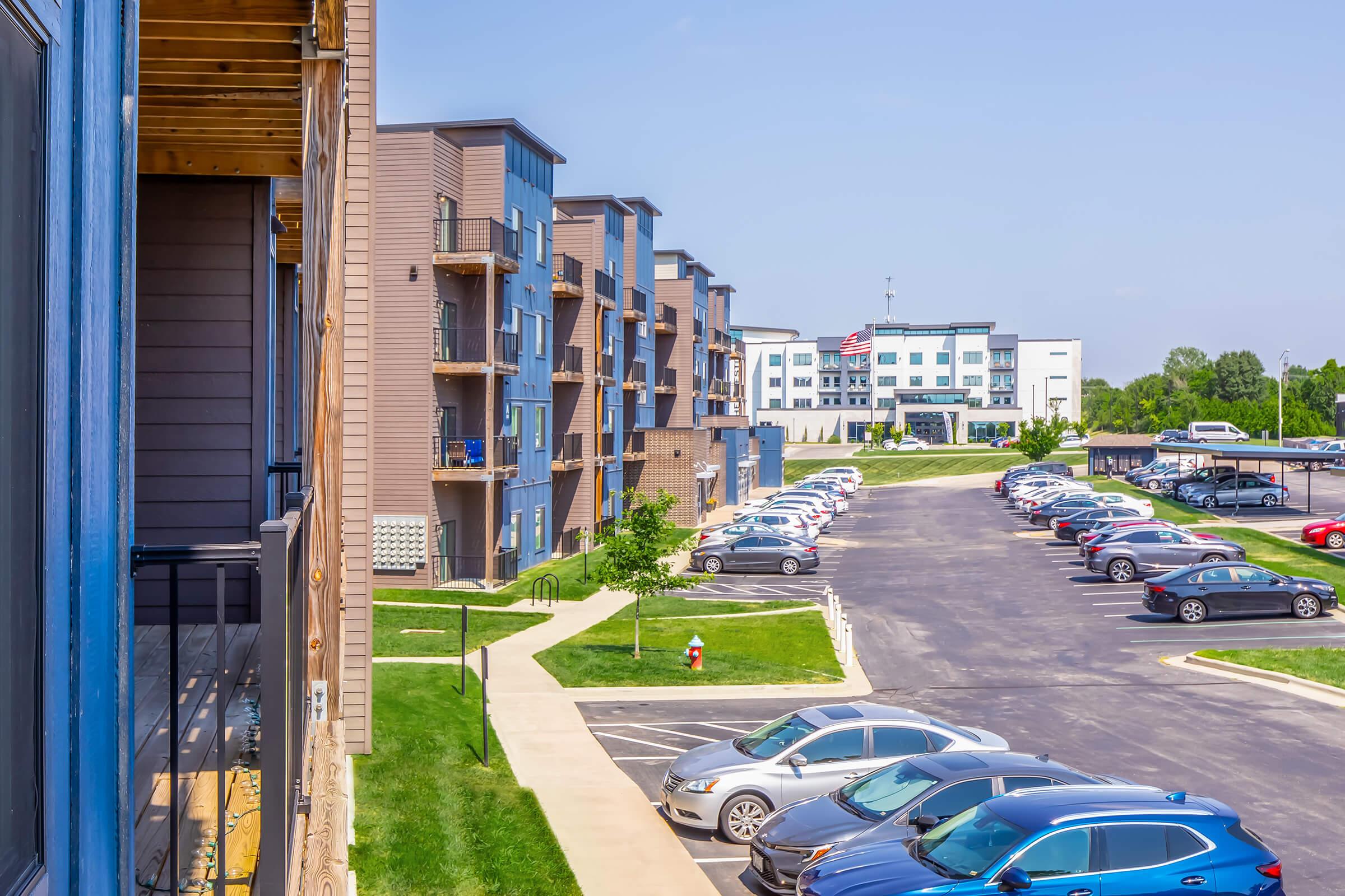 A view of an apartment complex with modern architecture, featuring multiple buildings and parked cars along a paved lot. Green lawns and landscaped areas are visible, under a clear blue sky. In the background, another hotel-like building can be seen with an American flag.