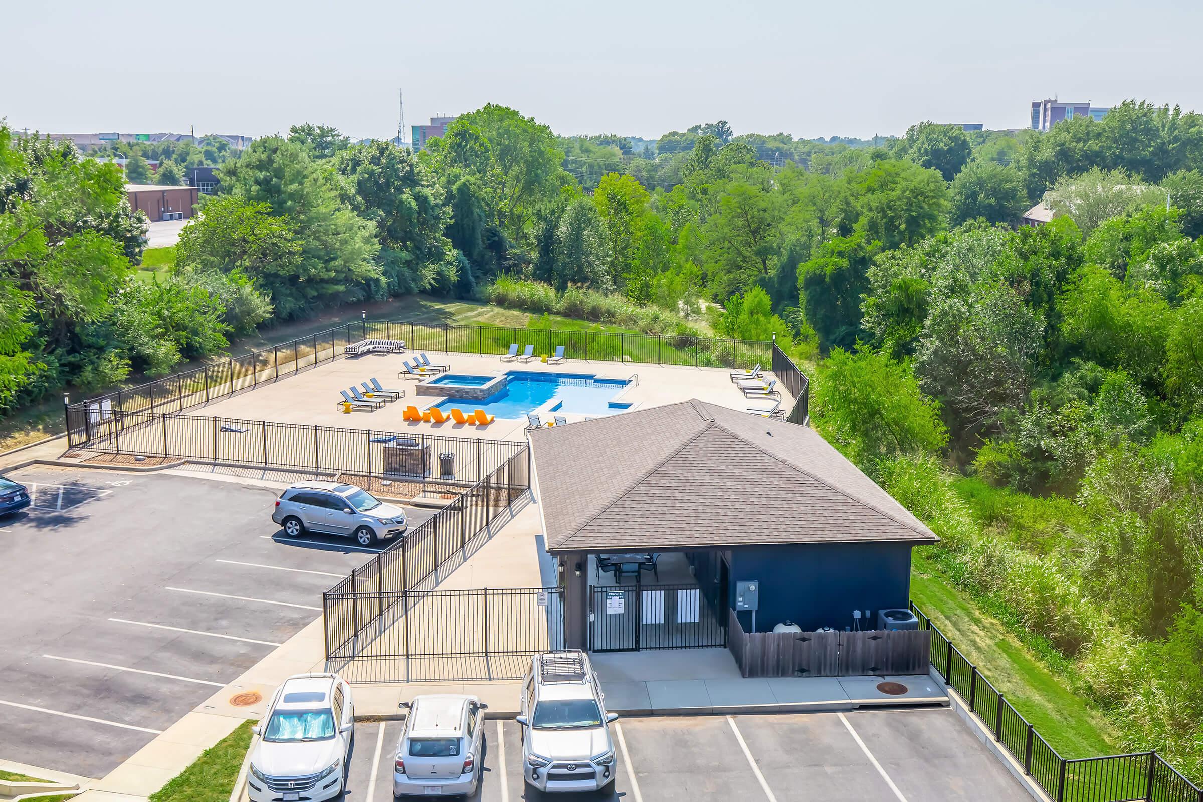 Aerial view of a community pool area featuring a fenced swimming pool with lounge chairs and umbrellas. A clubhouse is visible nearby, along with a parking lot filled with cars. Surrounding greenery adds to the tranquil setting.
