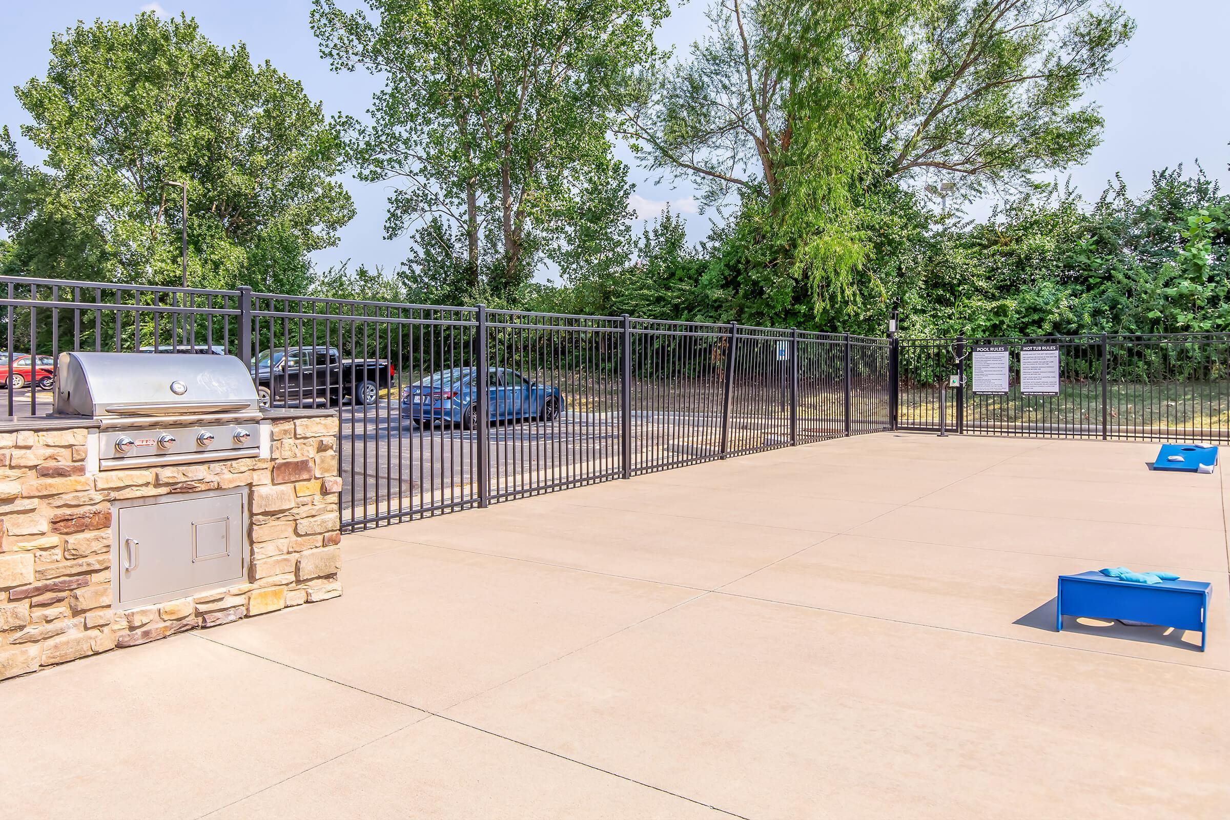 A concrete patio area featuring a stone-accented grill and two blue cornhole boards. The space is enclosed by a black fence and surrounded by trees and vegetation, with a parking area visible in the background. The setting appears to be suitable for outdoor gatherings or recreational activities.