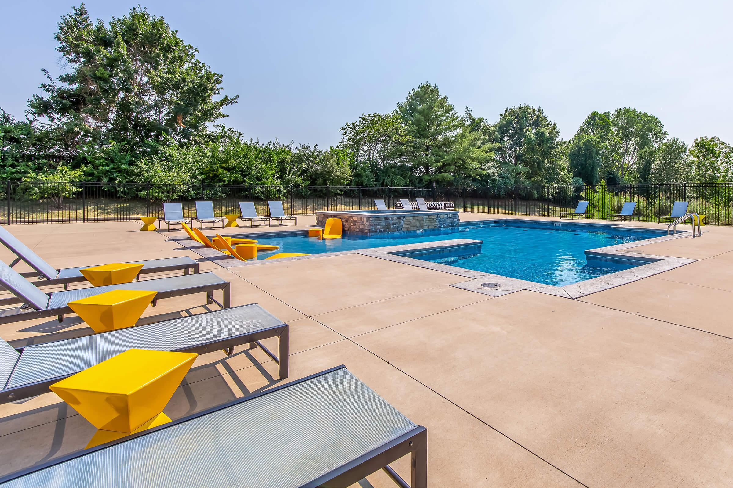 A sunny pool area featuring a blue swimming pool surrounded by concrete. There are several yellow lounge chairs and tables, with additional lounge chairs lined up nearby. Lush green trees provide a backdrop, and a section of the pool includes a shallow entry area.