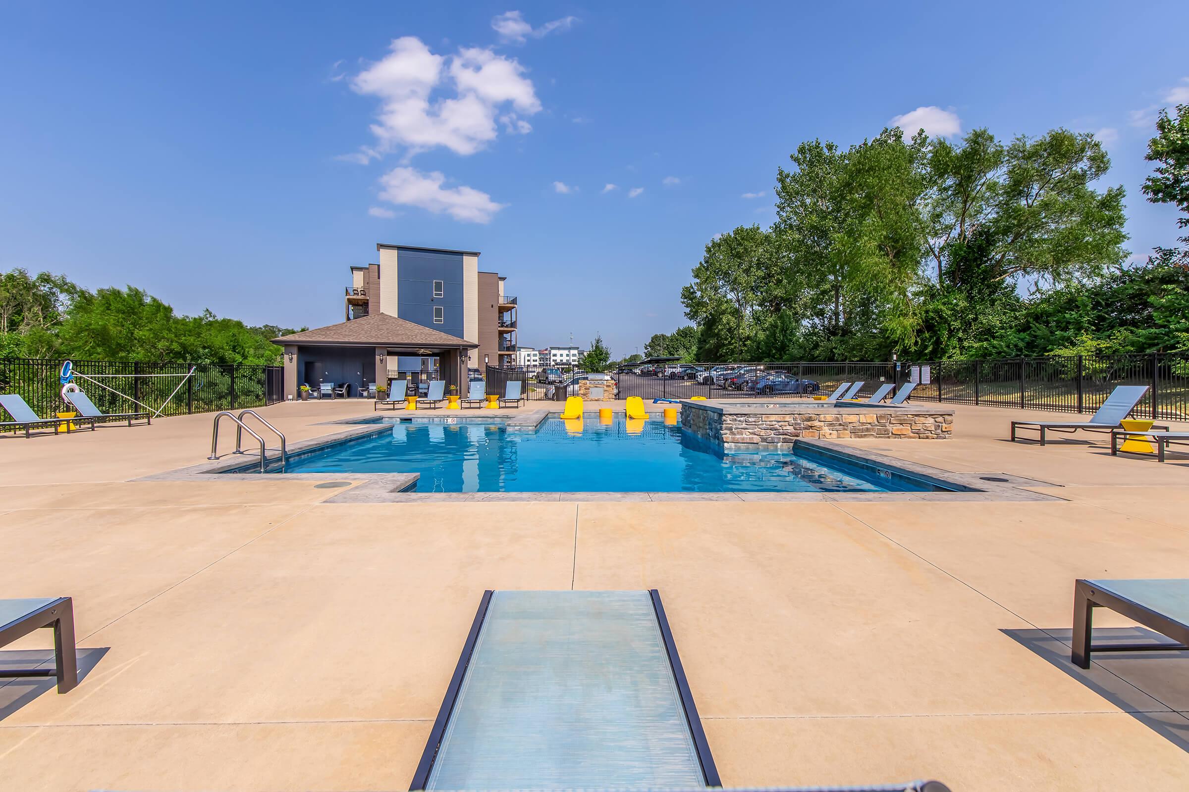 A sunny outdoor swimming pool area featuring bright yellow lounge chairs around the pool, surrounded by trees and a fence. In the background, there are modern buildings and a clear blue sky with a few clouds. The pool deck has several loungers positioned for relaxation.