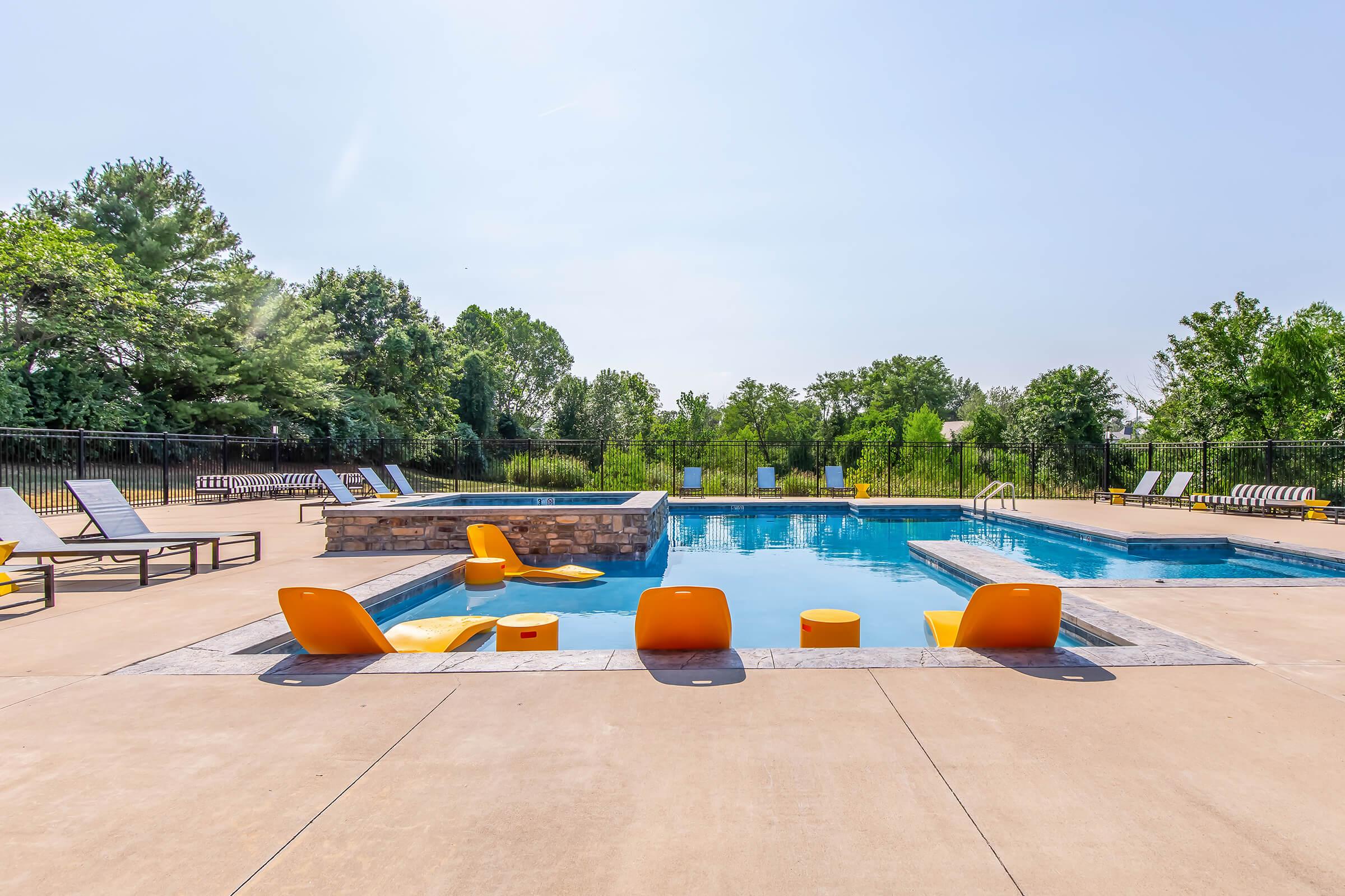 A bright, sunlit outdoor pool area featuring a clear blue pool with modern orange seating around it. Lounge chairs are arranged around the pool deck, surrounded by lush green trees in the background, creating a relaxing atmosphere. The scene is inviting and perfect for leisure activities.