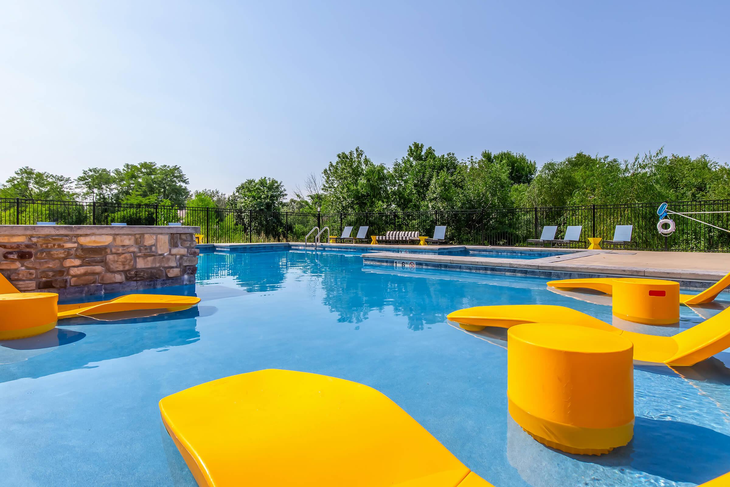 A sunny outdoor pool scene featuring clear blue water, with bright yellow lounge chairs scattered around. Lush green trees are visible in the background, and a light stone wall borders the pool area. The sky is clear and blue, creating a vibrant and inviting atmosphere.