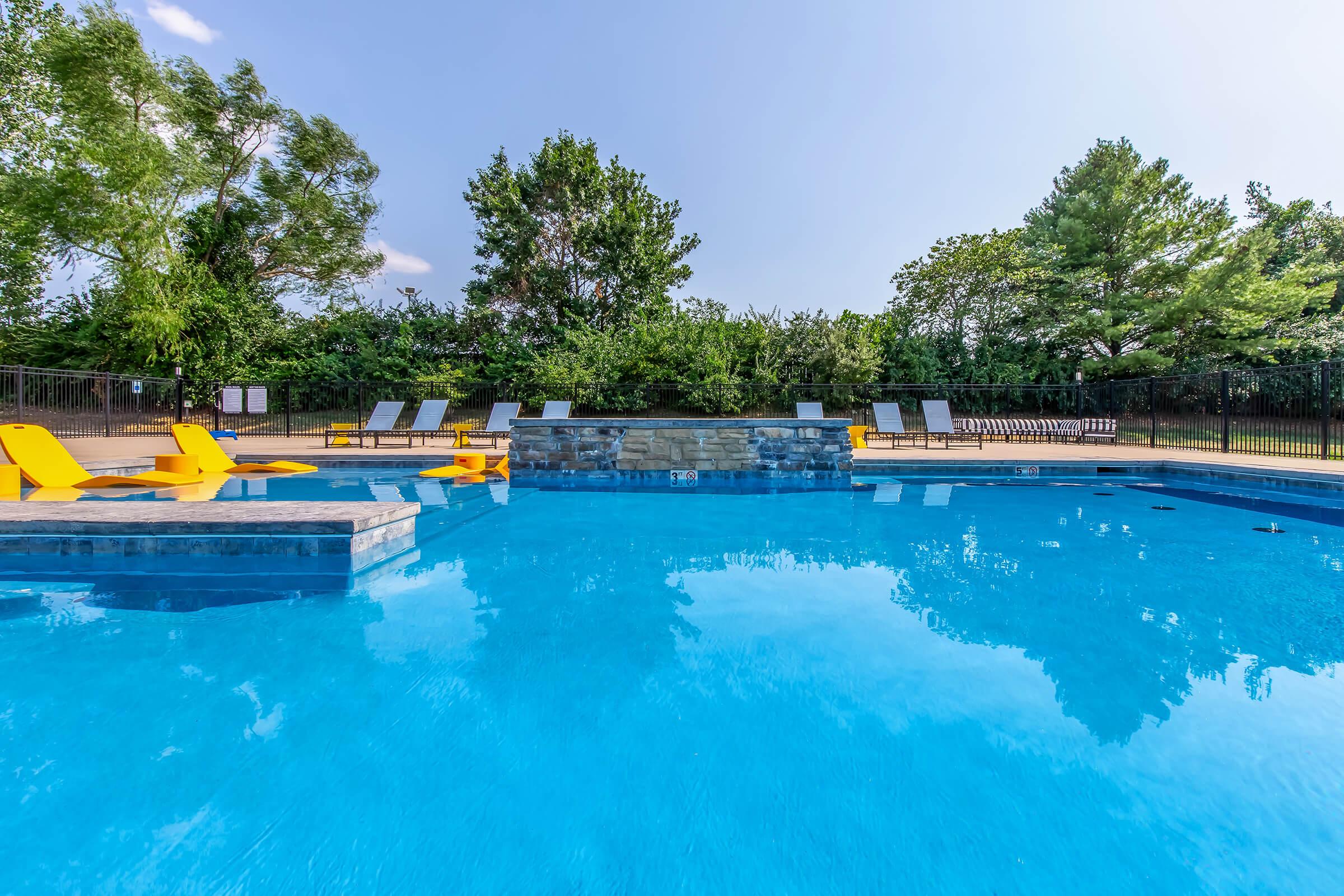 A serene swimming pool surrounded by lush greenery. The clear blue water reflects the sky, with several bright yellow lounge chairs arranged on the pool deck. In the background, trees provide shade, and a fenced area enhances the private setting. Ideal for relaxation and leisure.
