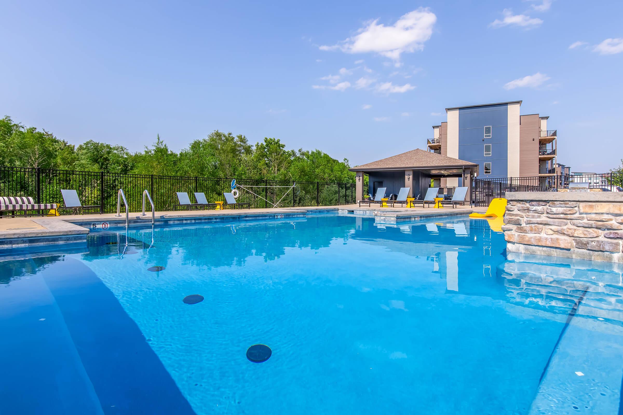 A clear blue swimming pool surrounded by lounge chairs and a stone patio. In the background, a building with multiple floors is visible, along with greenery. The sky is bright with a few clouds, creating a welcoming and relaxing atmosphere.