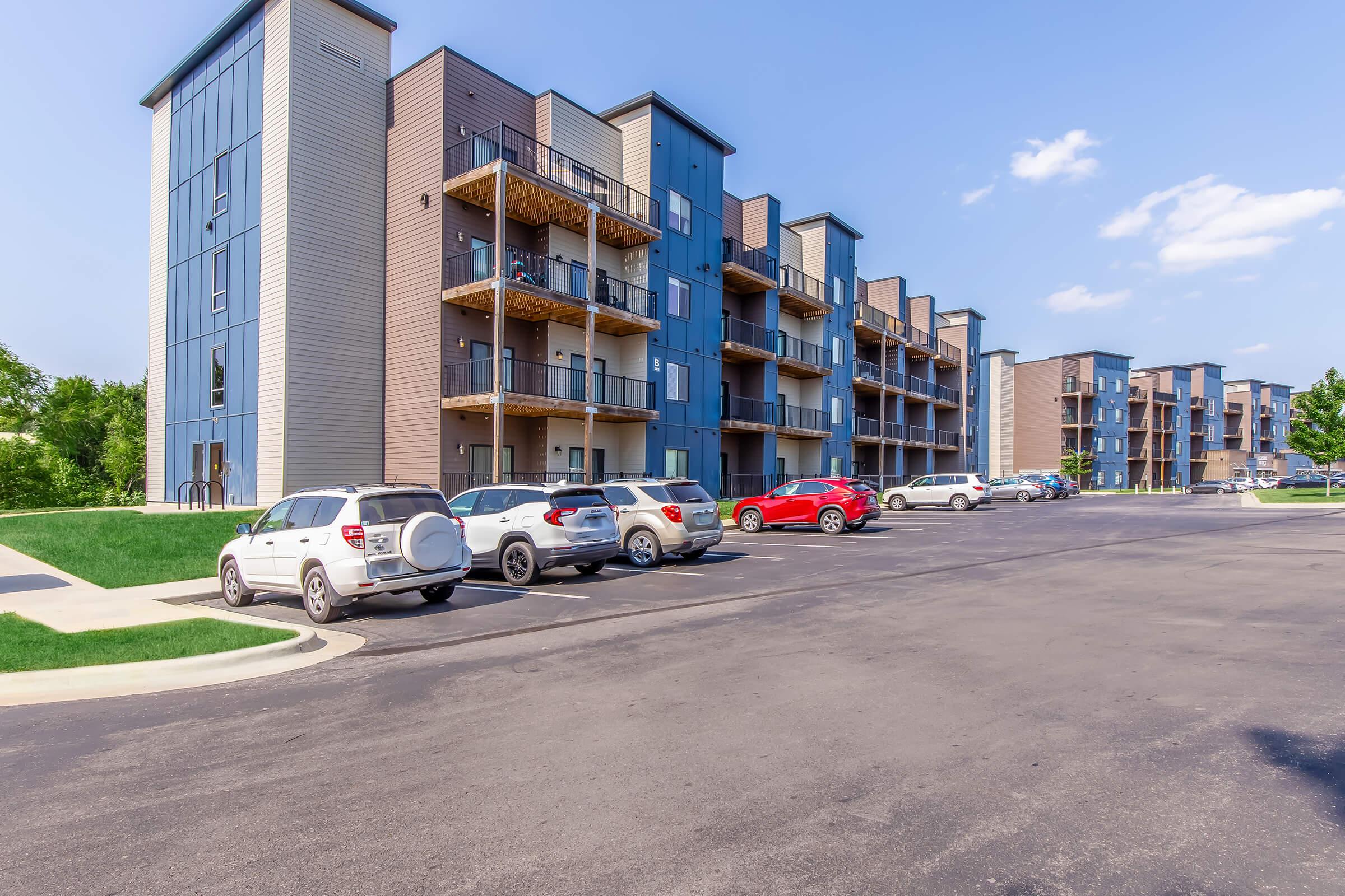 Modern apartment buildings with multiple floors, featuring balconies. A parking lot in the foreground has several parked cars, including white SUVs and a red sedan. Green grass and trees are visible, along with a clear blue sky and a few clouds.