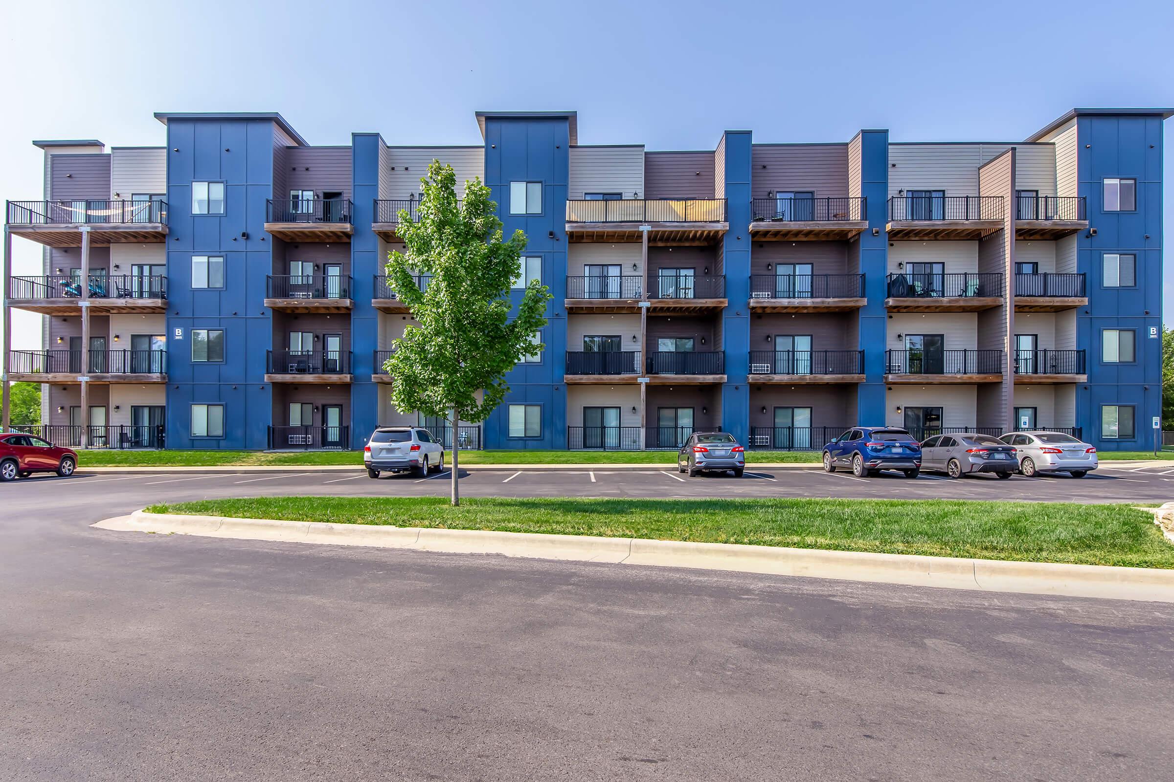 A modern, multi-story blue apartment building featuring multiple balconies. The building is surrounded by a landscaped area with green grass and a paved parking lot. Several cars are parked in front, and clear blue skies are visible above.
