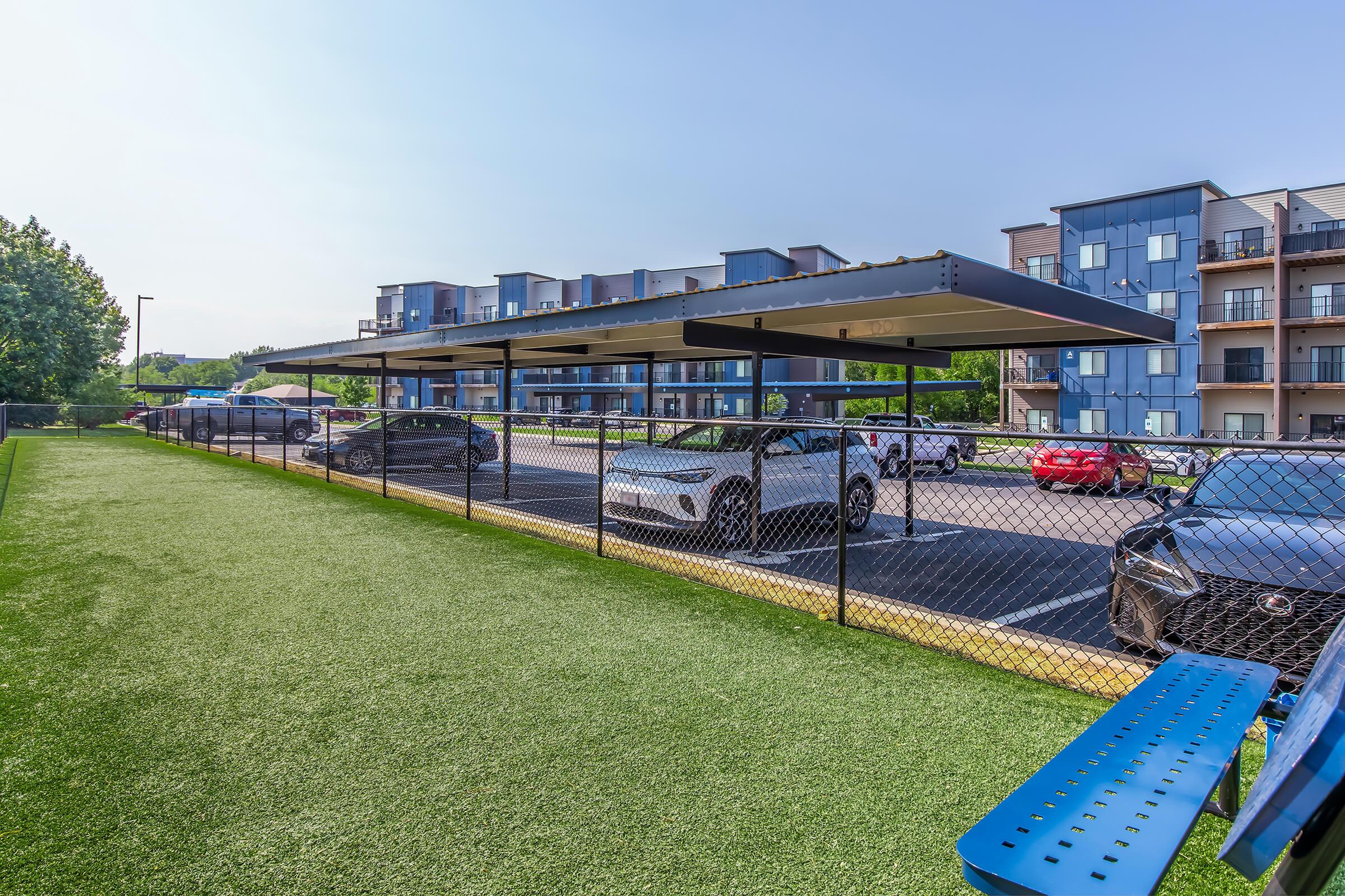 A parking area featuring covered spaces for vehicles, surrounded by a chain-link fence. In the background, modern apartment buildings are visible, along with a green grass area in the foreground. Clear blue sky above creates a bright atmosphere.