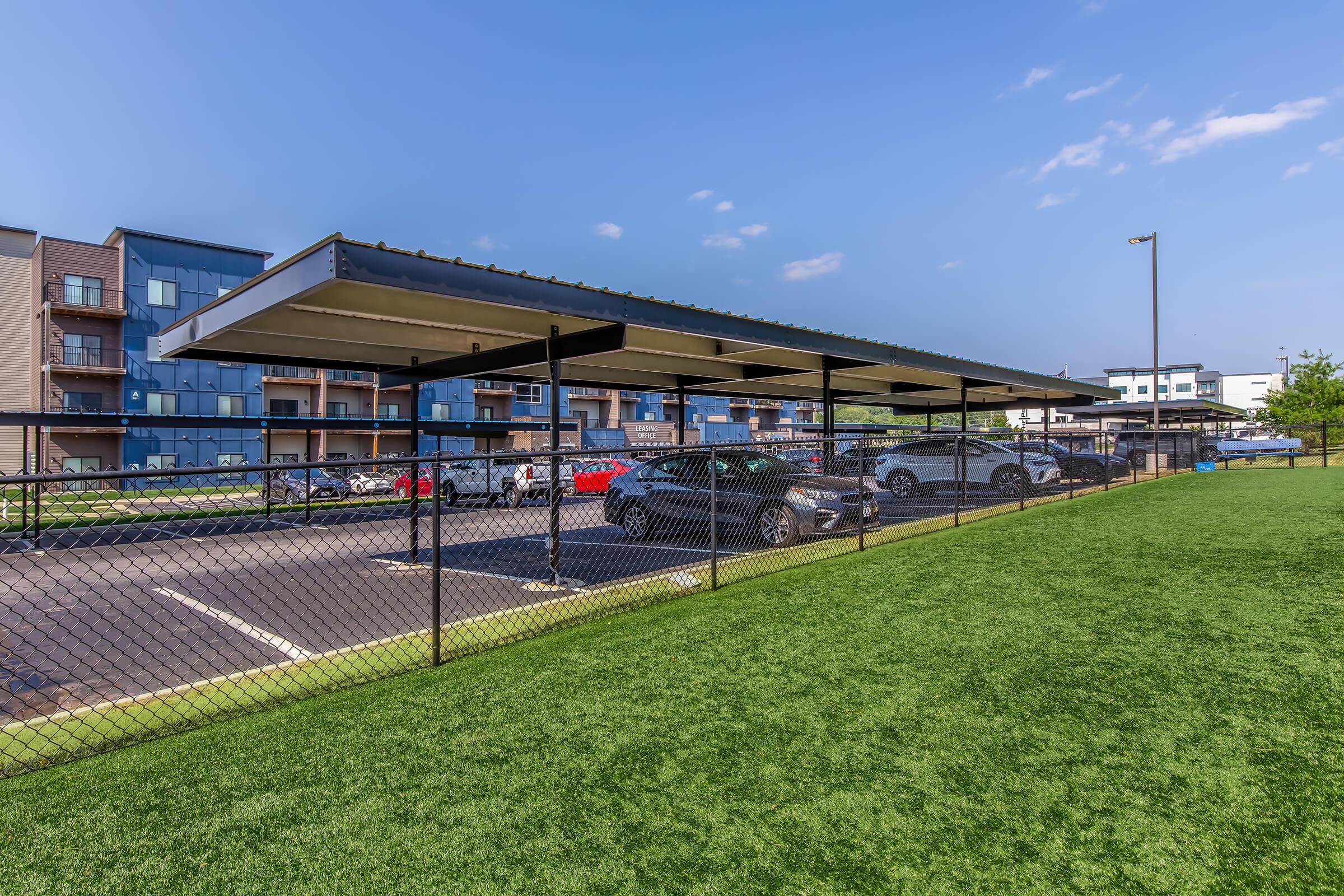 Carport area with several parked cars, surrounded by a black fence. In the background, there are modern residential buildings with balconies. The ground is covered in artificial grass, and the sky is clear with a few clouds.
