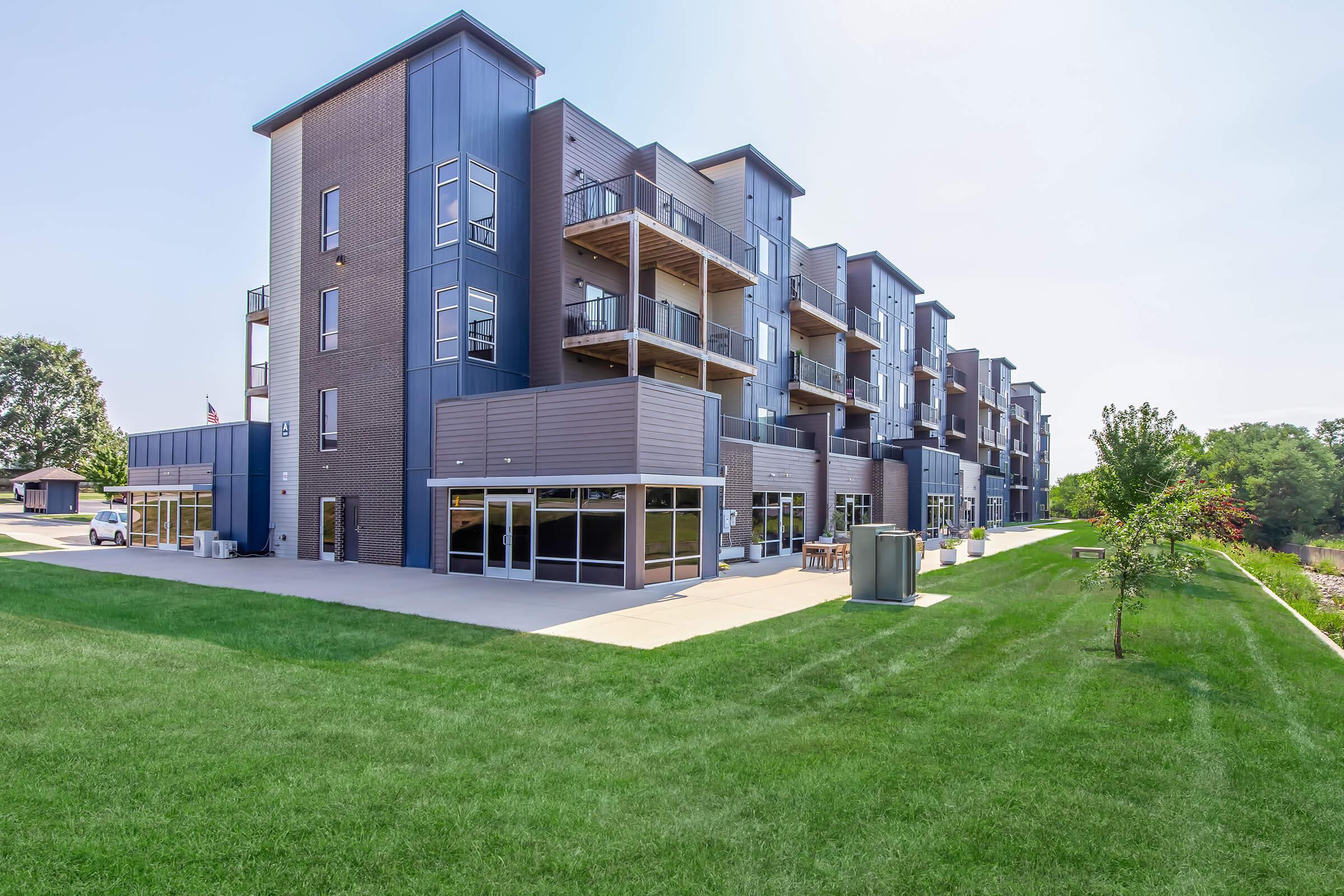 Modern apartment building with multiple stories featuring balconies. The exterior is a mix of gray brick and metal panels. In the foreground, there is a well-maintained green lawn and some trees. The area appears spacious and inviting, with large windows reflecting sunlight.