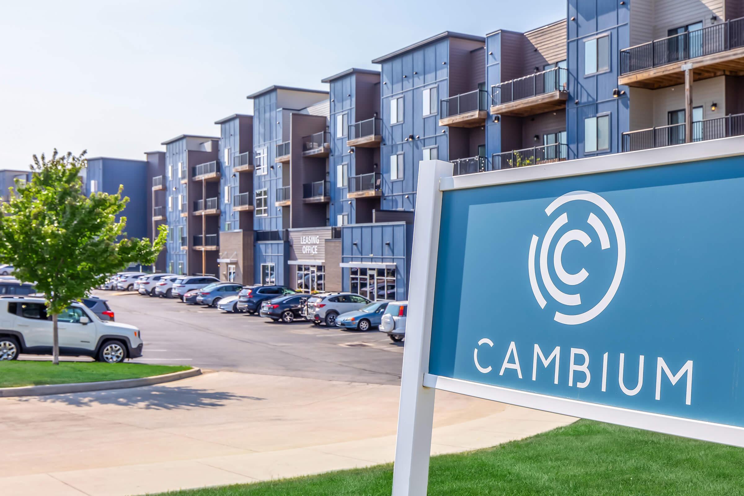 A modern apartment complex featuring multiple three- to four-story buildings with balconies. A blue sign in the foreground displays the name "CAMBIUM." The parking lot is filled with vehicles, and there are trees and green lawns around the area, suggesting a well-maintained residential environment.