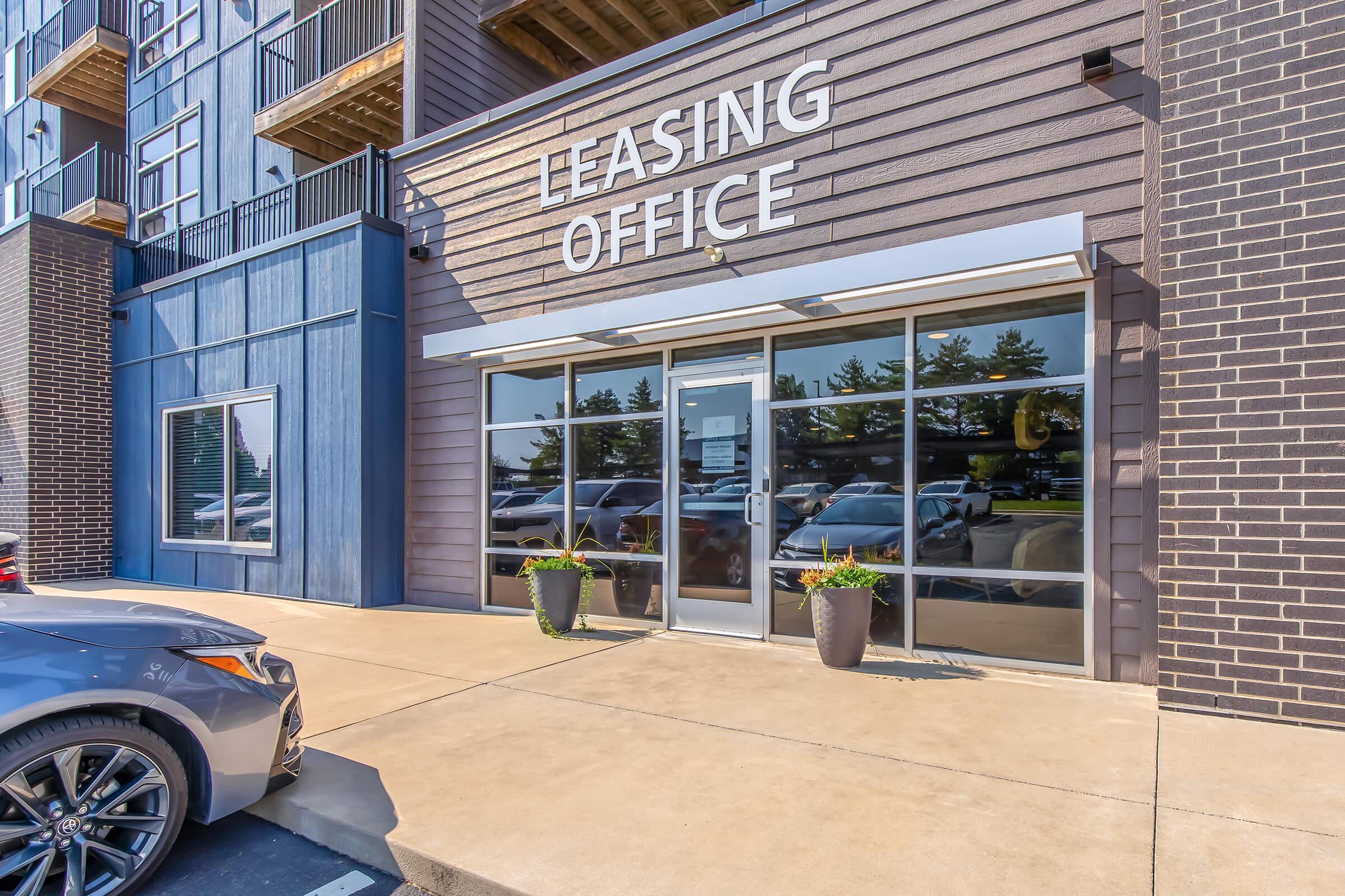 Exterior view of a leasing office with large glass doors. The building features a modern design with a dark exterior and prominent signage reading "Leasing Office." Planters with plants are positioned near the entrance. A parked car is visible in front of the office.