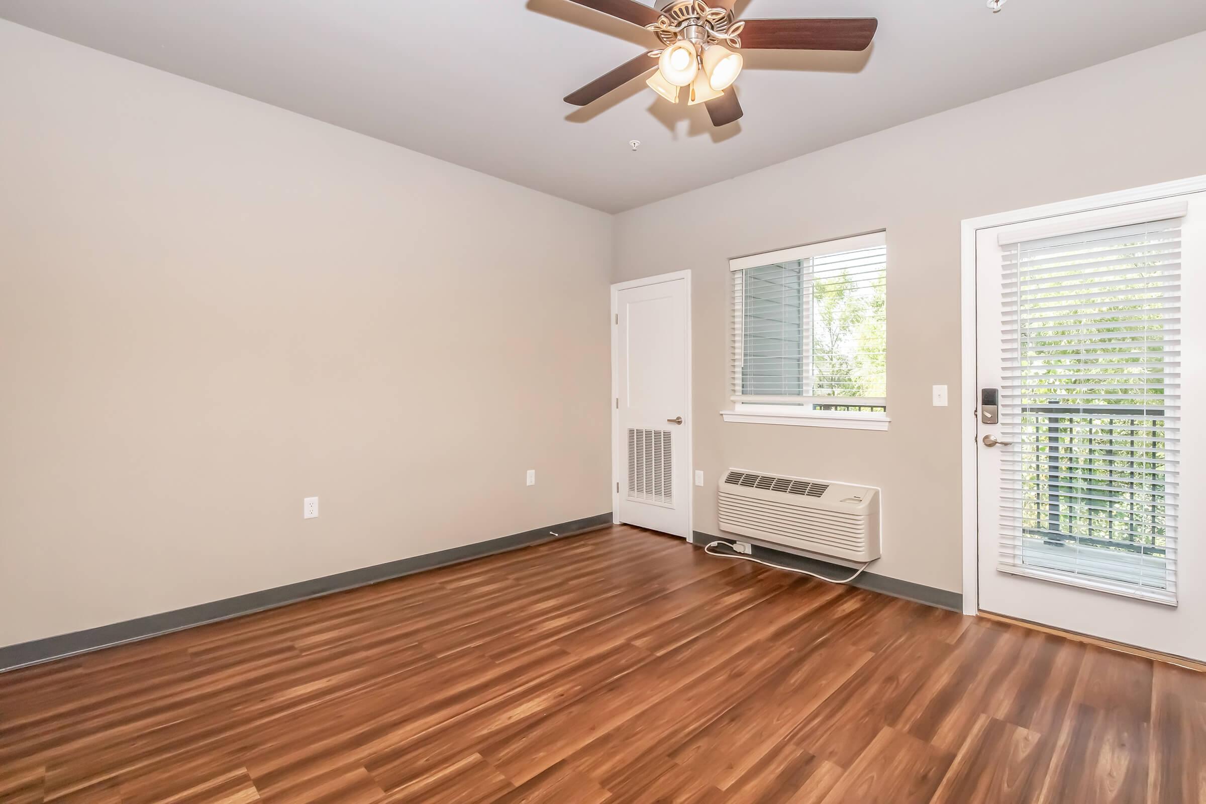 Interior view of a room featuring light gray walls, a ceiling fan with lights, and laminate wood flooring. A door with a window leads to a balcony, and there is an air conditioning unit on the wall. Natural light streams in through the window, creating a bright and open atmosphere.