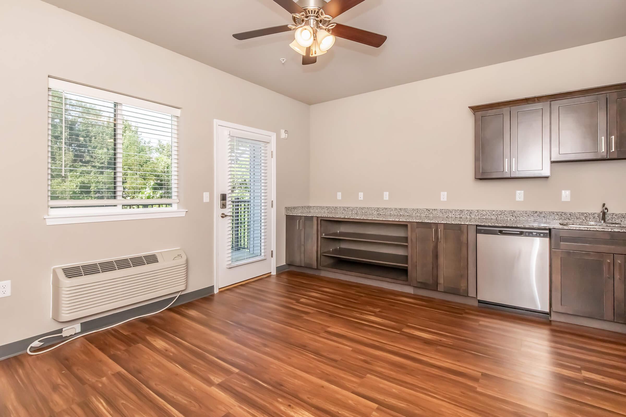 Interior of a modern kitchen and living area featuring hardwood flooring, stainless steel appliances, a granite countertop, and a ceiling fan. There is a window with blinds and a glass door leading to a balcony, providing natural light and a view of trees outside.