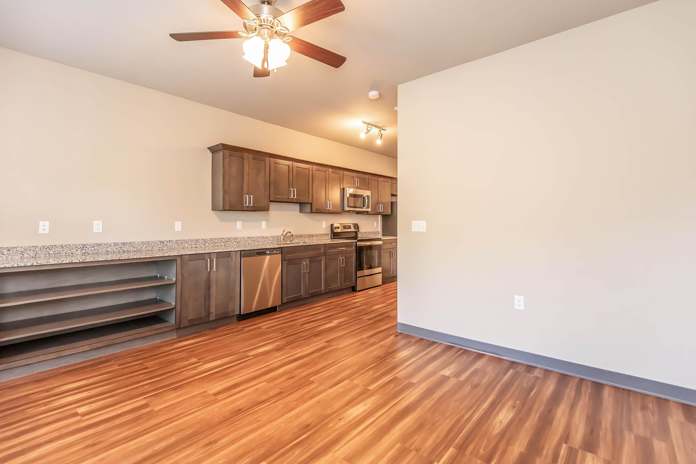 Modern kitchen and living area featuring dark wood cabinets, stainless steel appliances, and a ceiling fan. The flooring is a warm, wood-like laminate. The open layout includes a countertop space and shelves, creating a spacious and inviting atmosphere.