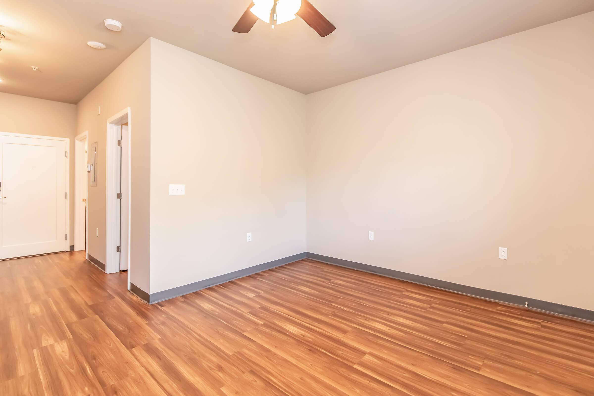 A spacious, empty room with light brown laminate flooring and beige walls. The ceiling features a ceiling fan. Doors are visible on the left and right walls, and there is a doorway leading to the entrance on the far left. The overall ambiance is bright and inviting, with natural light enhancing the space.