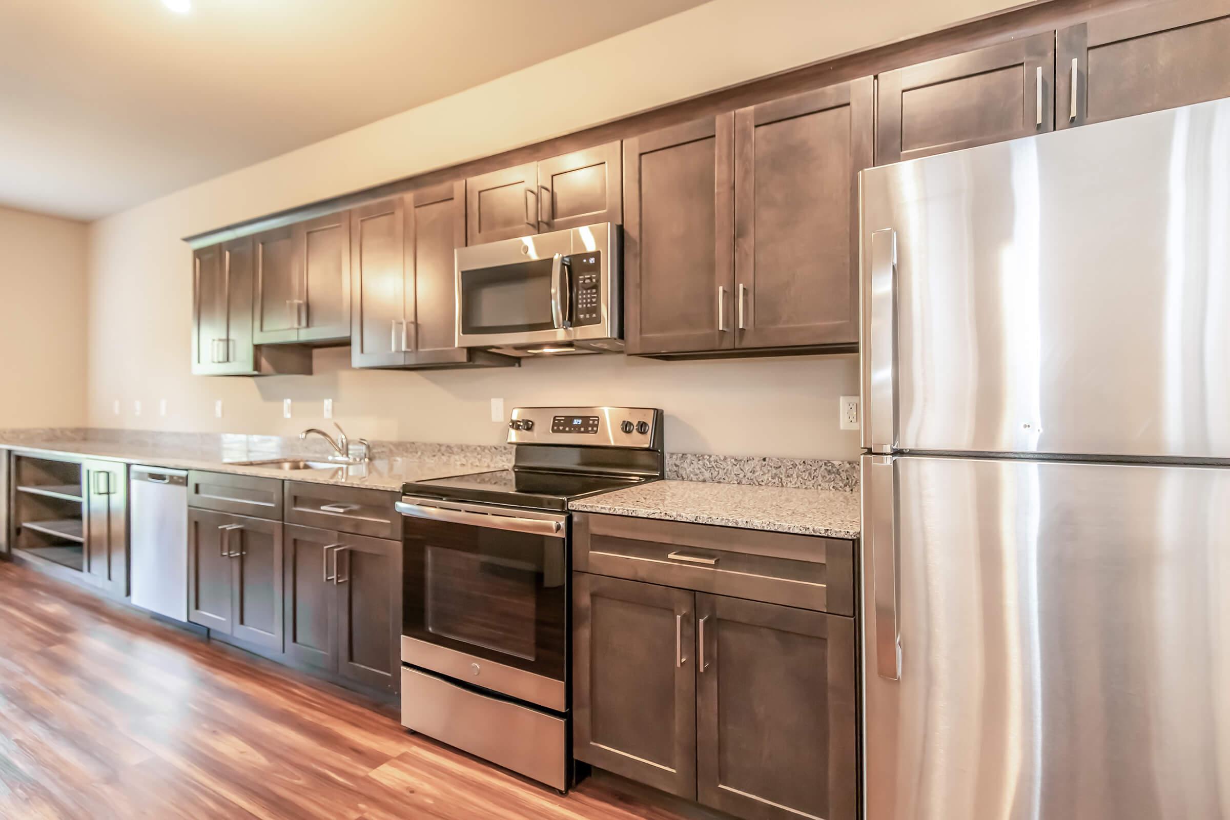 Modern kitchen featuring dark wood cabinetry, stainless steel appliances including an oven, microwave, and refrigerator. The countertops are made of granite, and the flooring is a warm wood finish. Ample storage is provided by cabinets and shelves. The space is well-lit with natural and overhead lighting.