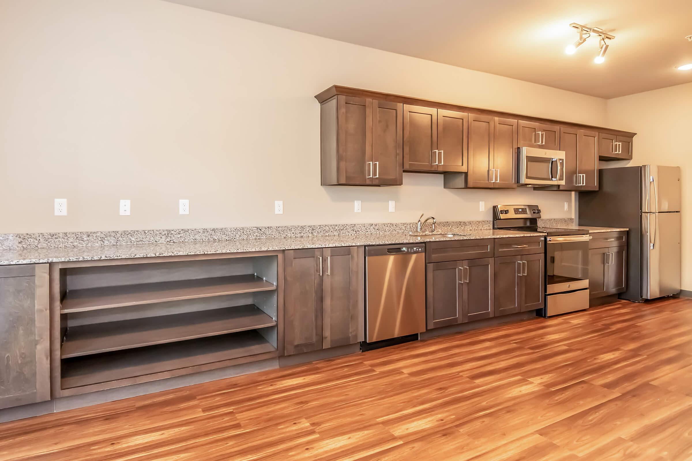 Modern kitchen featuring dark wood cabinetry, stainless steel appliances including an oven, microwave, dishwasher, and refrigerator. The kitchen has a granite countertop, with open shelving beneath and hardwood floors. Bright lighting enhances the spacious appearance.