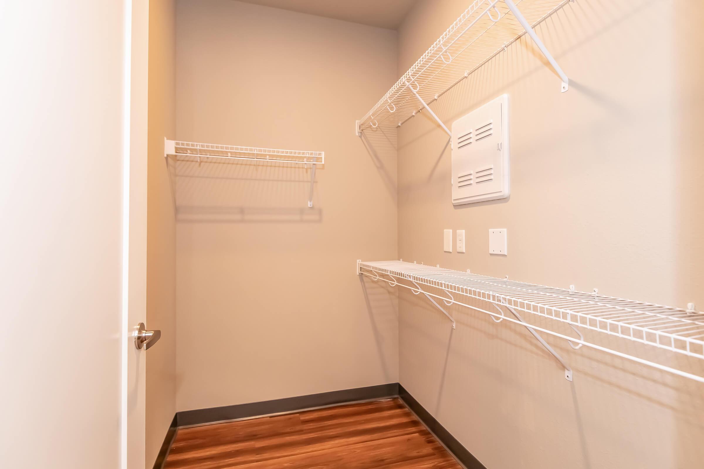 A small, empty closet with beige walls and a wooden floor. There are two wire shelving units mounted on the walls, one on the left side and another running along the back wall. The closet door is partially open, revealing the interior space. There is a white vent on the wall and two electrical outlets visible.