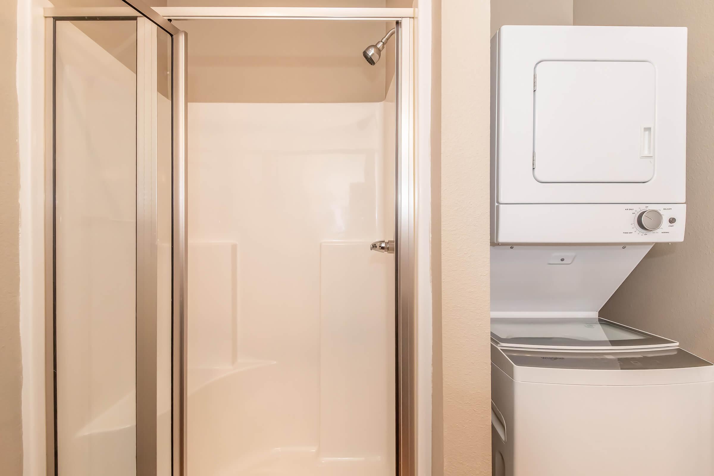 A clean bathroom corner featuring a glass-enclosed shower stall next to a stacked washer and dryer unit. The walls are a neutral color, enhancing the bright and tidy appearance of the space.