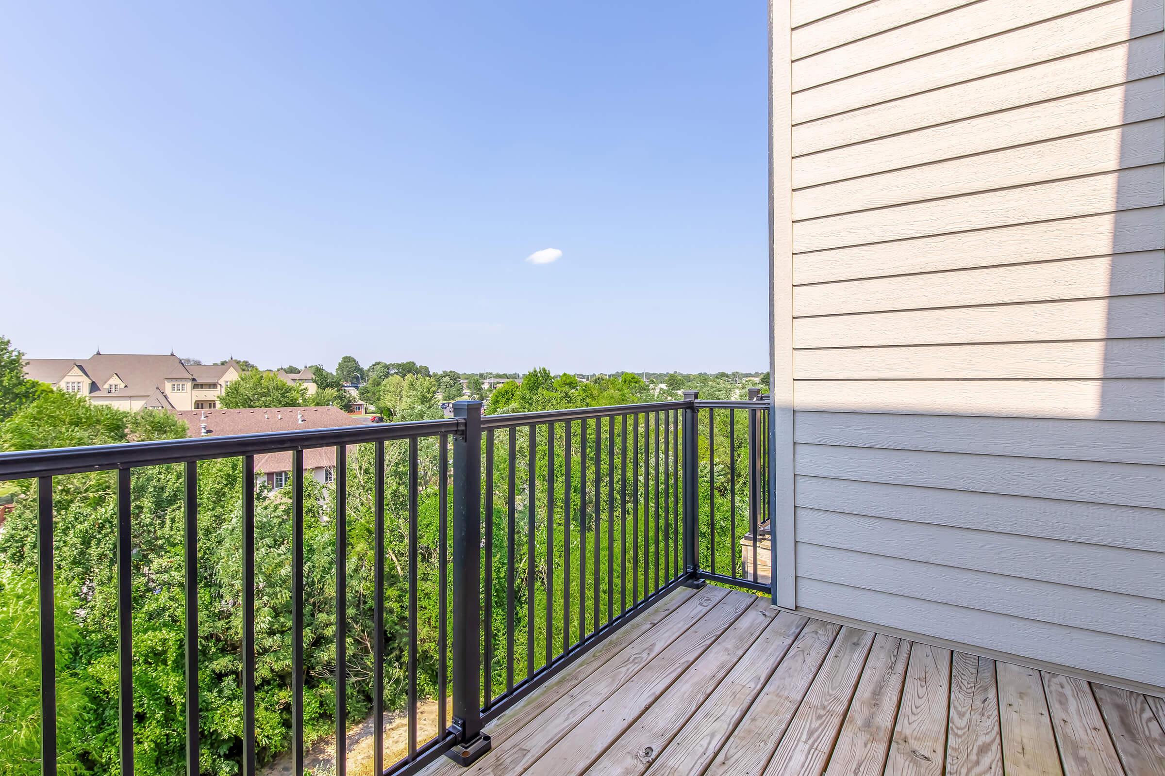 View from a balcony overlooking a green landscape with trees and distant houses under a clear blue sky. The image features a railing made of black metal and wooden flooring, giving a sense of spaciousness and tranquility.