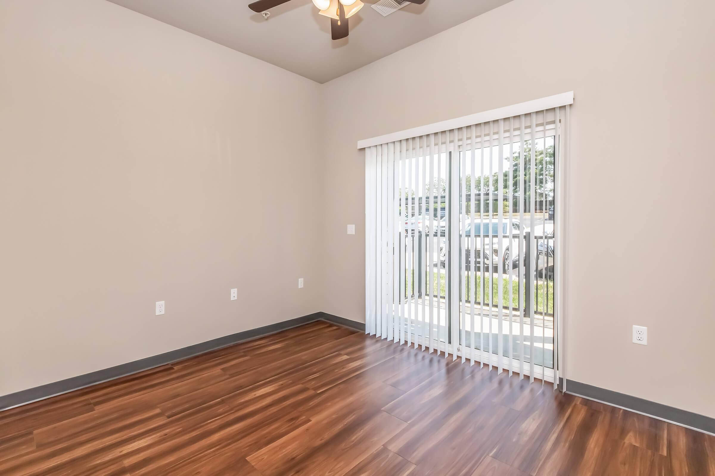 A spacious room with light brown wooden flooring, neutral-colored walls, and a ceiling fan. There is a large sliding glass door with vertical blinds leading to an outdoor area, allowing natural light to enter. The room is empty, creating a clean and minimalistic atmosphere.