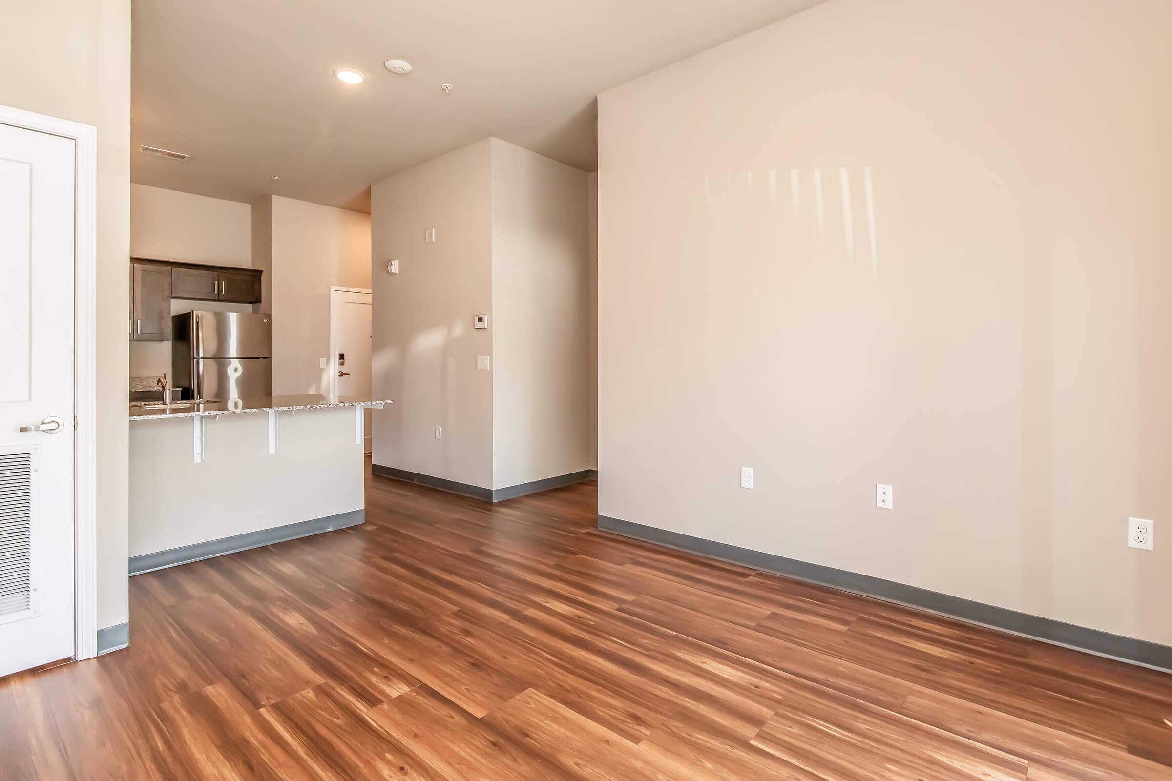An empty modern apartment interior featuring hardwood floors, light-colored walls, and an open layout. The kitchen area is partially visible with stainless steel appliances, and there is natural light streaming in from a nearby window. The space is clean and uncluttered, ready for furnishings.