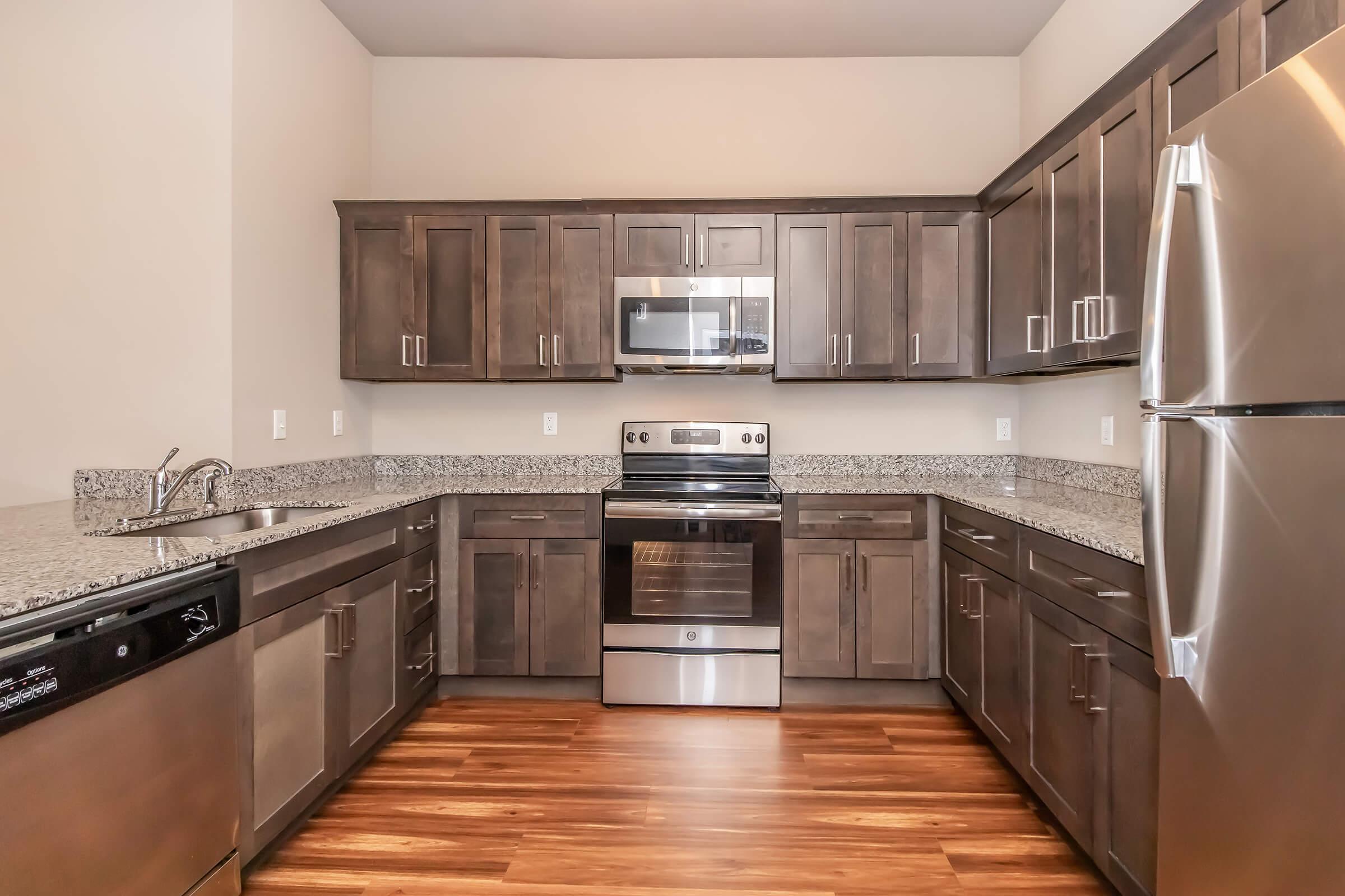 Modern kitchen featuring dark wooden cabinets, stainless steel appliances, granite countertops, and a sleek design. The kitchen includes a refrigerator, oven, microwave, and a dishwasher, with ample storage and workspace, complemented by hardwood flooring.