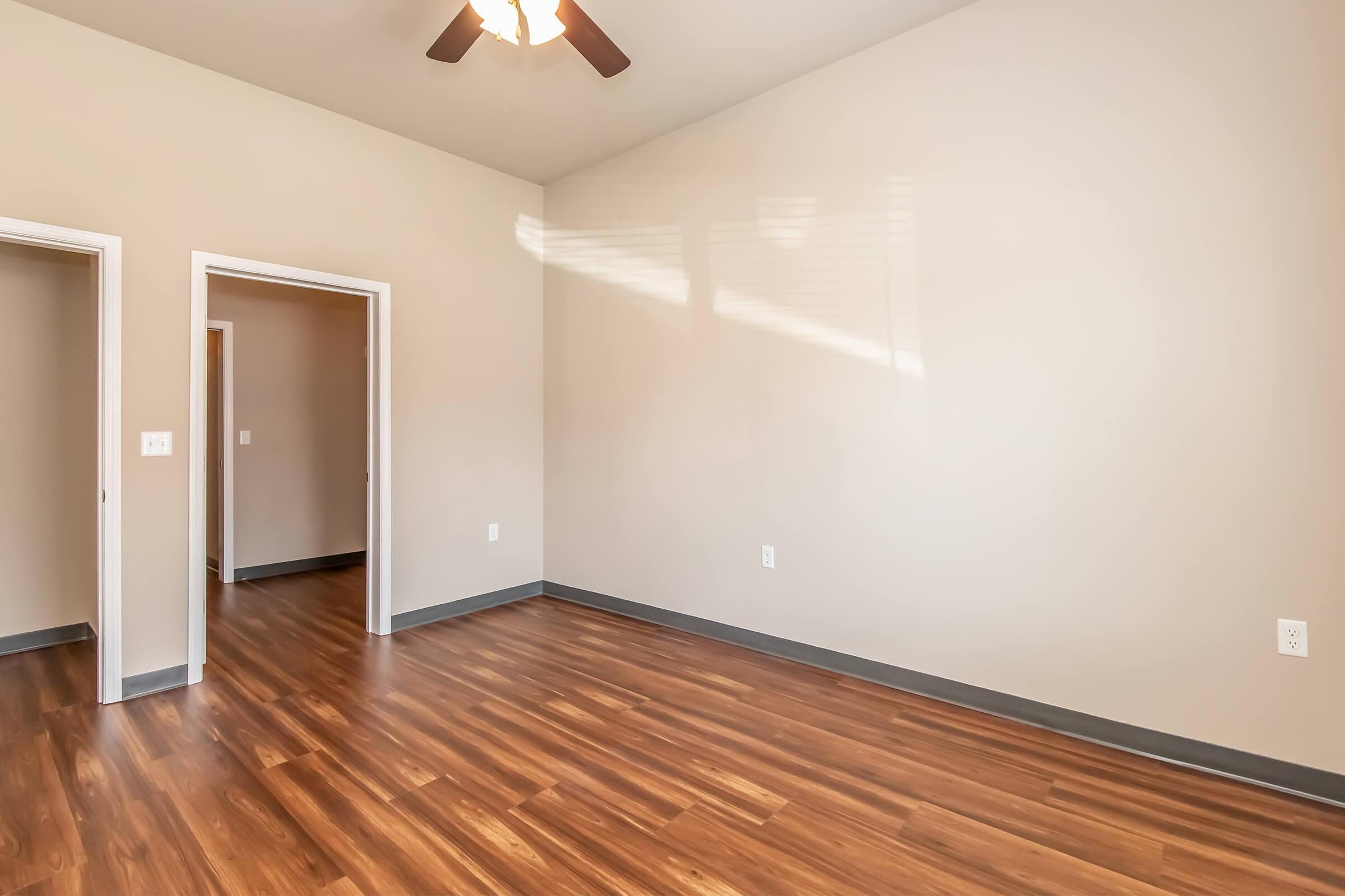 An empty room with light brown wooden flooring and beige walls. There are two doorways leading to other rooms, and a ceiling fan is visible on the ceiling. Natural light is streaming through a window, casting shadows on the floor. The space is clean and uncluttered, creating an open feel.
