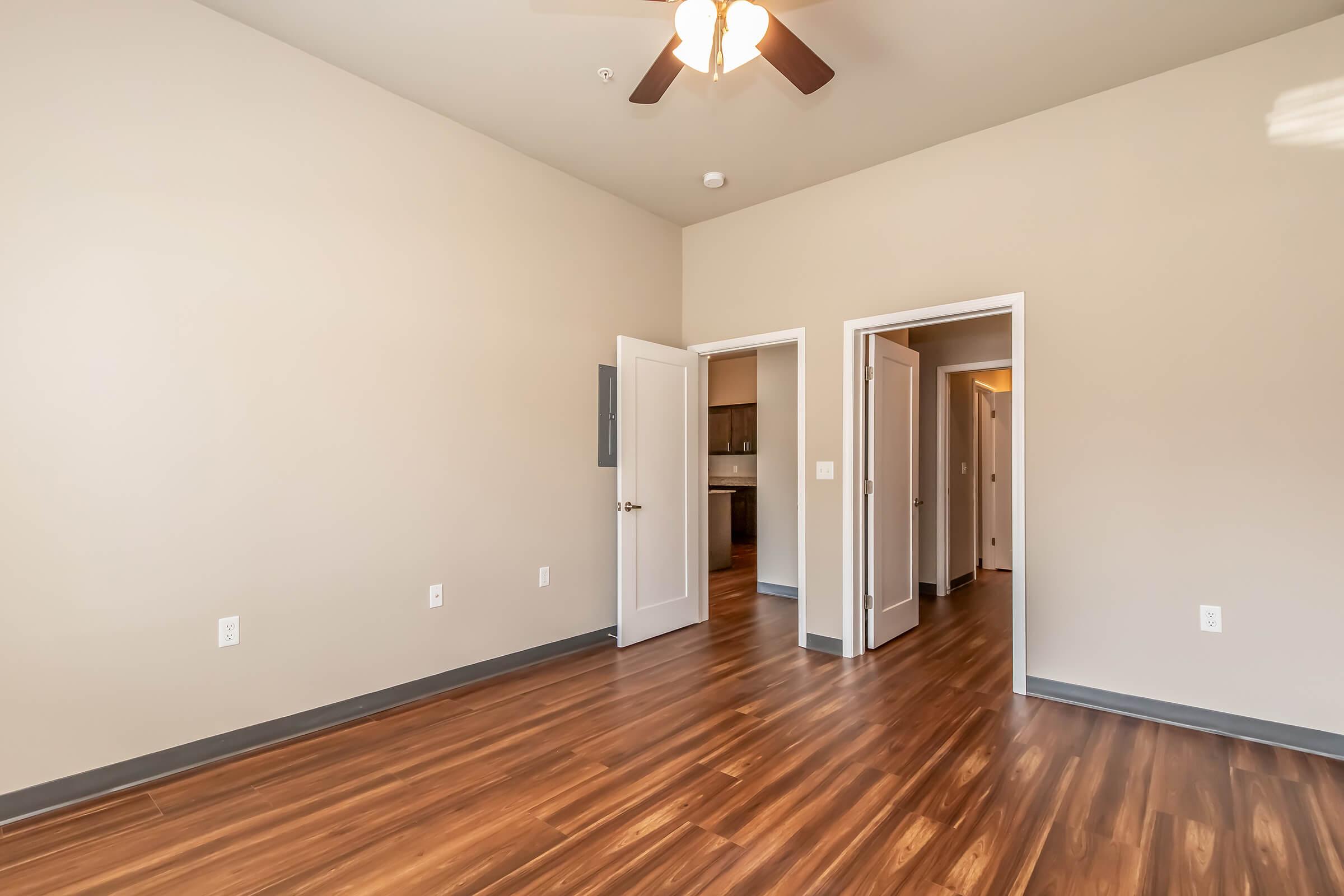 A spacious room with light-colored walls and a wooden floor, featuring a ceiling fan. Two white doors lead to adjoining areas, and a window brings in natural light. The room has a simple, modern design.