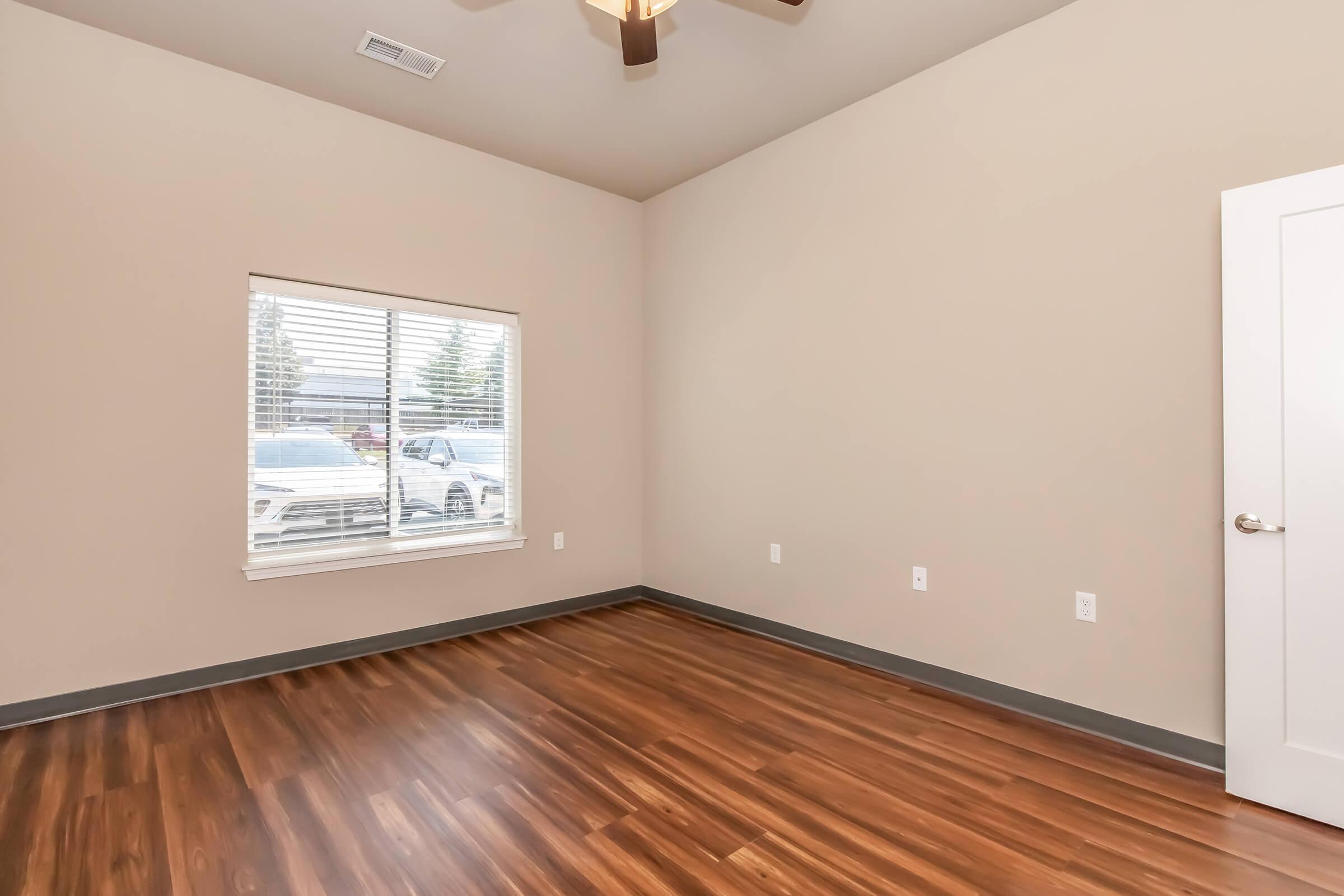 A spacious, empty room with light beige walls and a wooden floor. It features a ceiling fan and a window with white blinds, allowing natural light to enter. Outside the window, parked cars are visible. The overall ambiance is clean and ready for furnishing.