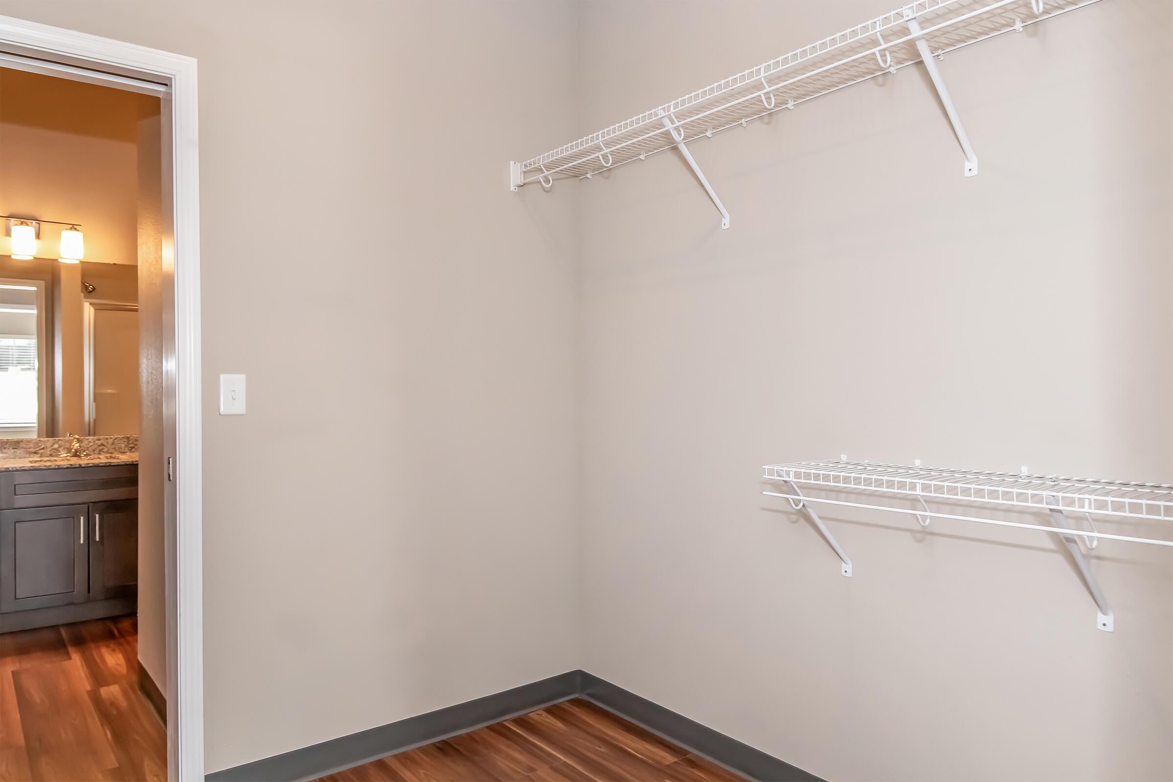 An empty closet with two wire shelving units mounted on a beige wall. The floor has wooden laminate, and there is an open doorway leading to a bathroom area with vanity lights visible in the background.