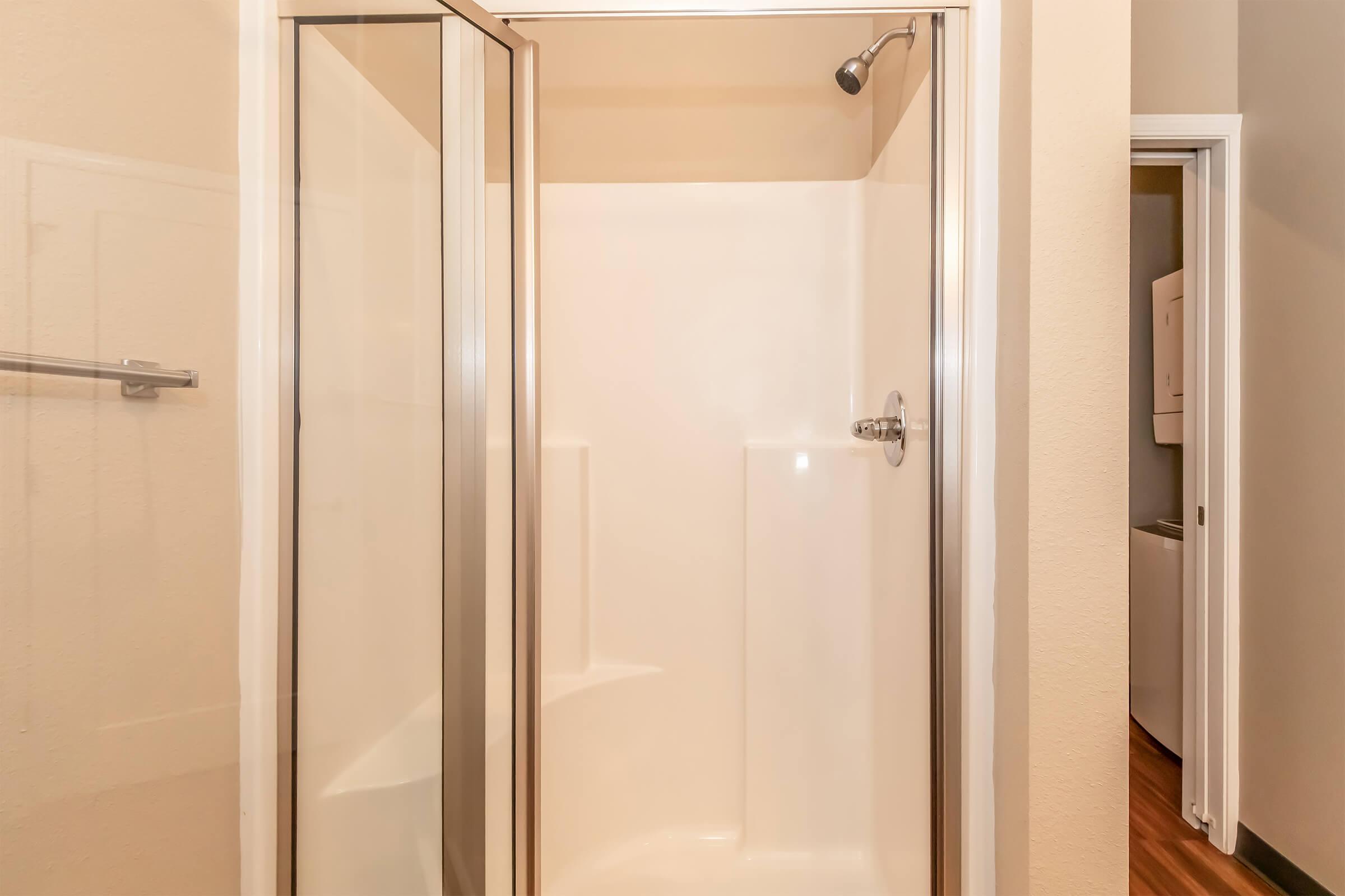 A clean, empty glass shower enclosure with a chrome showerhead. The walls are light-colored, and the floor is wooden. A doorway leads to another room, providing a glimpse of the interior layout.