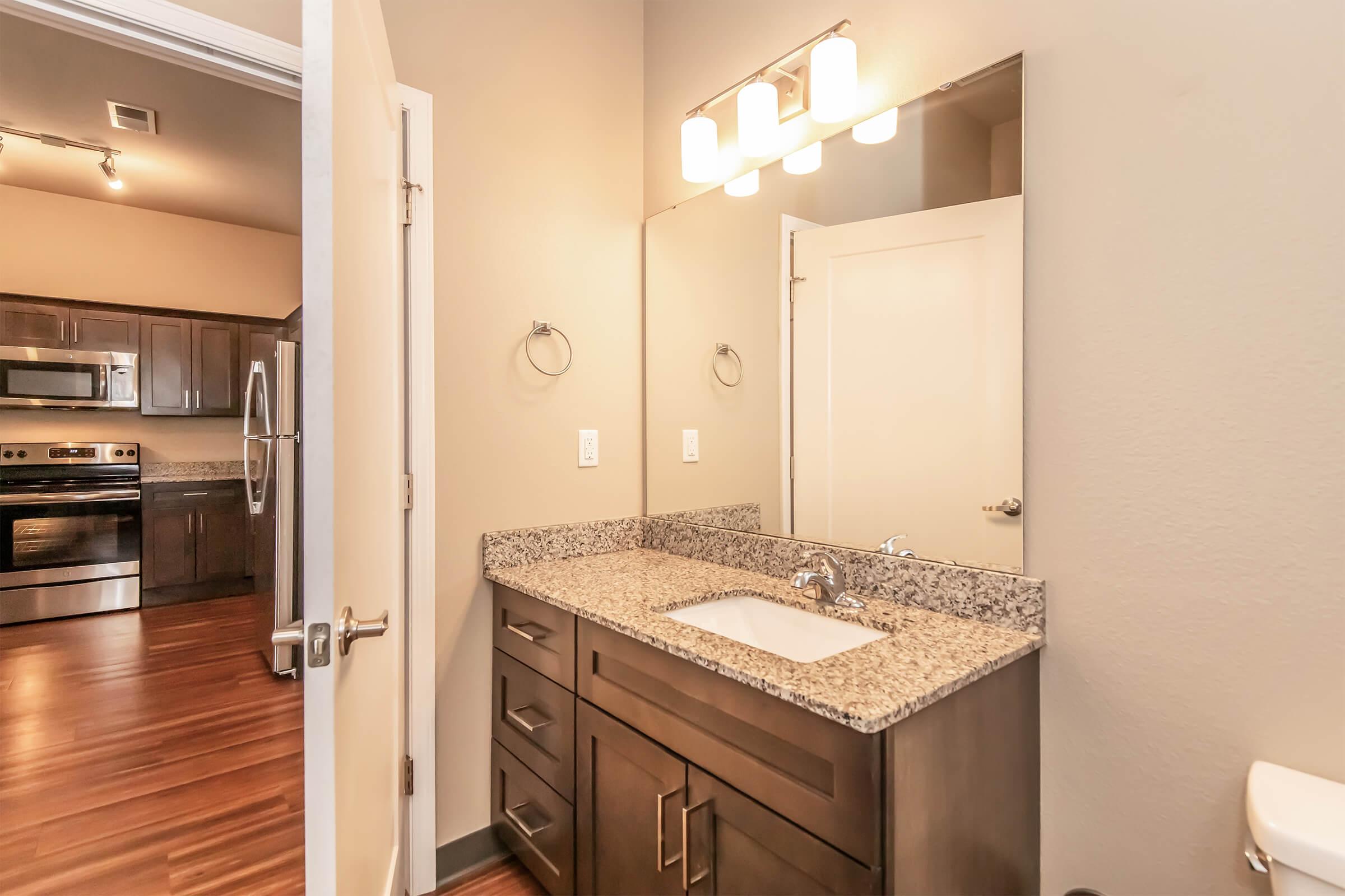 A modern bathroom featuring a granite countertop with a sink and a large mirror above. The walls are painted a neutral color, and there's a hand towel ring on the side. The doorway leads into a kitchen area with dark wood cabinets and stainless steel appliances visible in the background.