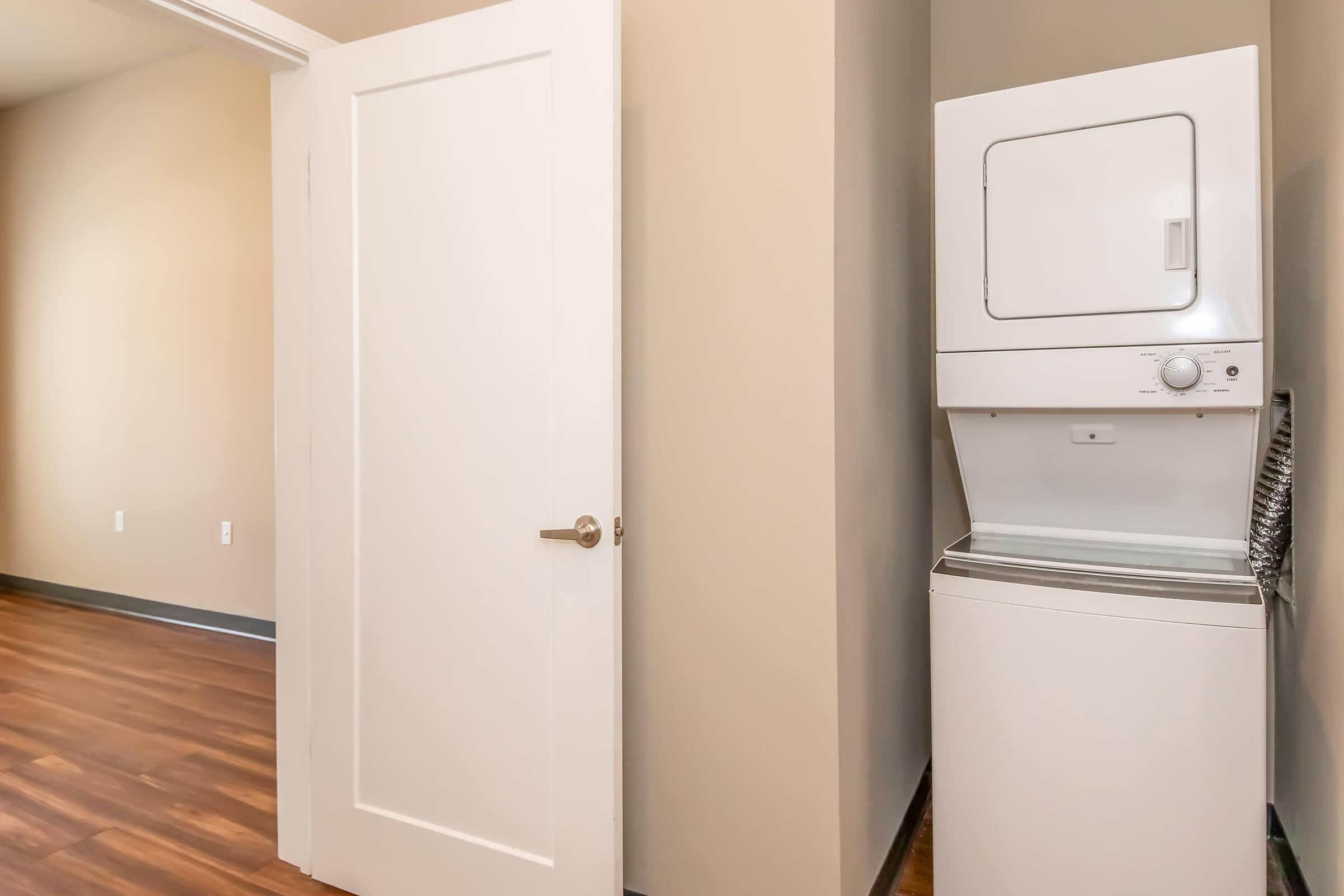 A compact laundry area featuring a stacked washer and dryer against a light beige wall, with a white door leading to another room. The floor is wooden, and the space has a modern, minimalist design.