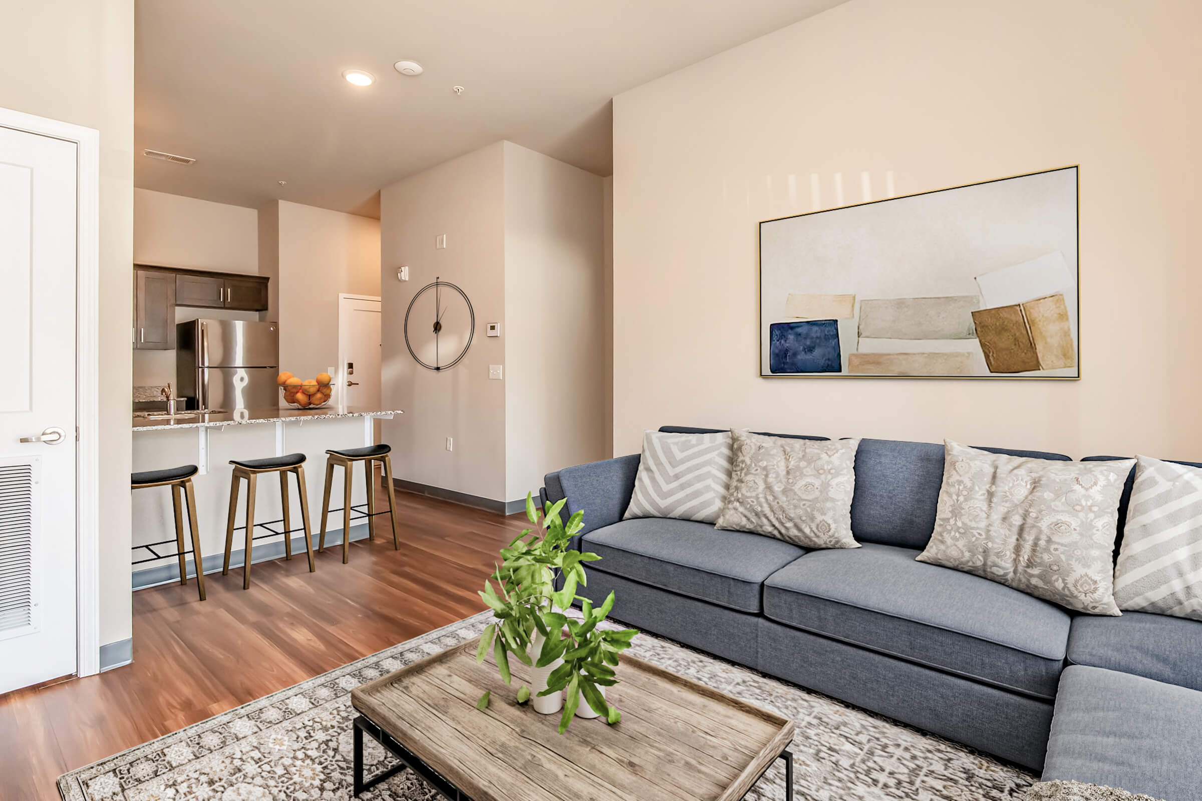A modern living room featuring a spacious gray sofa with decorative pillows, a patterned rug, and a small green plant on a coffee table. In the background, a kitchen with bar stools can be seen, along with a piece of abstract art hanging on the wall. The space has warm tones and natural light.