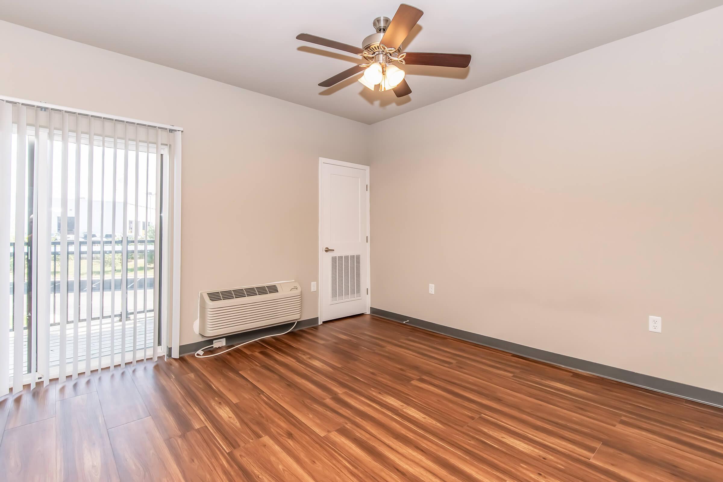 An empty room featuring a ceiling fan with wooden blades, light brown walls, and hardwood flooring. There is a large window with vertical blinds allowing natural light and an air conditioning unit on the wall. A white door is visible, leading to another space.