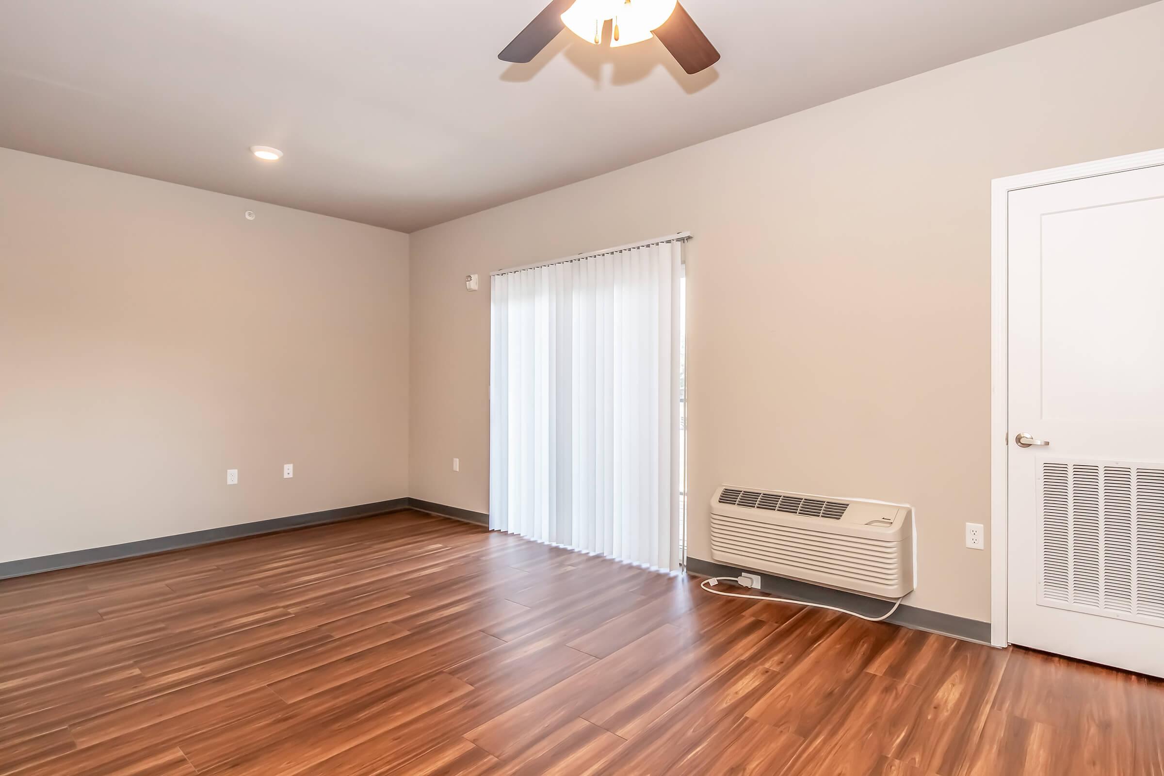 Spacious interior of a room featuring wood laminate flooring, a ceiling fan, a wall-mounted air conditioner, and a large window with white sheer curtains. The walls are painted in a neutral color, and there is a plain door on the right. The overall aesthetic is modern and inviting.