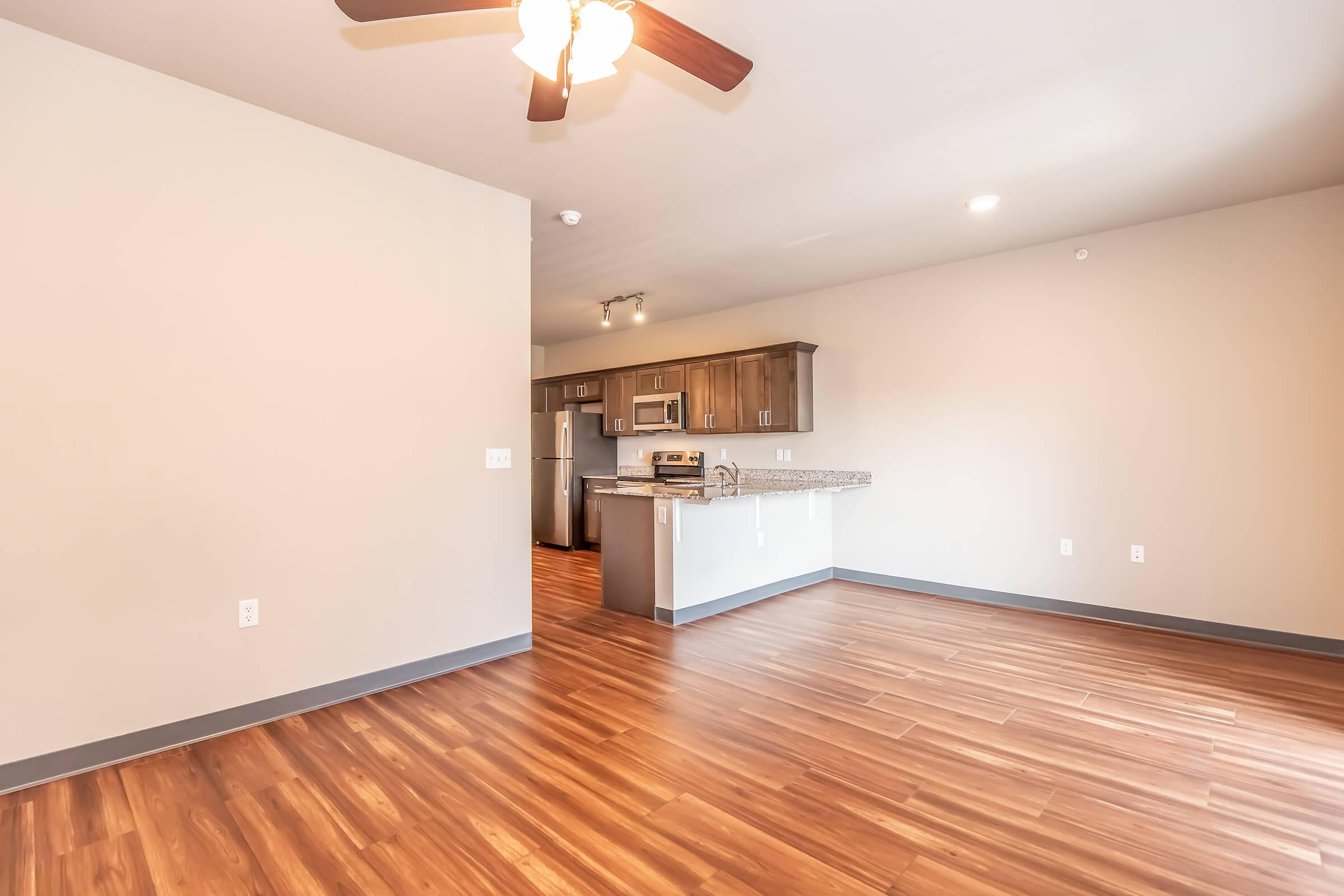 Interior view of a modern apartment showcasing a living area with hardwood-style flooring, a ceiling fan, and a kitchen area in the background featuring stainless steel appliances and wooden cabinetry. The walls are painted in a neutral color, enhancing the spacious feel of the room.