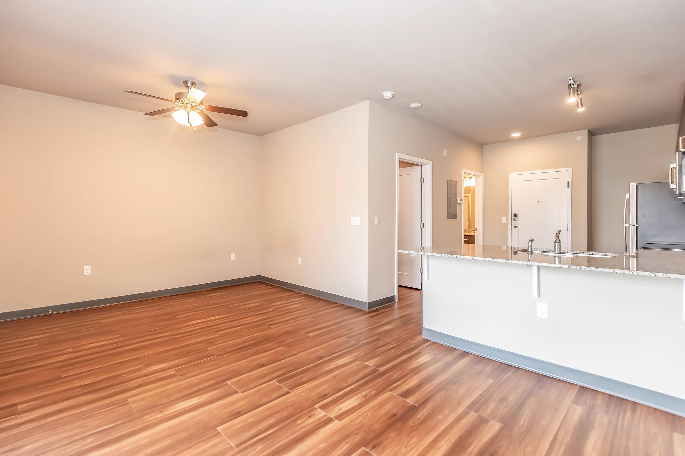 Spacious living area featuring hardwood-style flooring, a ceiling fan, and an open layout. A modern kitchen with a breakfast bar is visible in the background, along with doorways leading to other rooms. The walls are painted in a neutral color, enhancing the light and airy atmosphere.