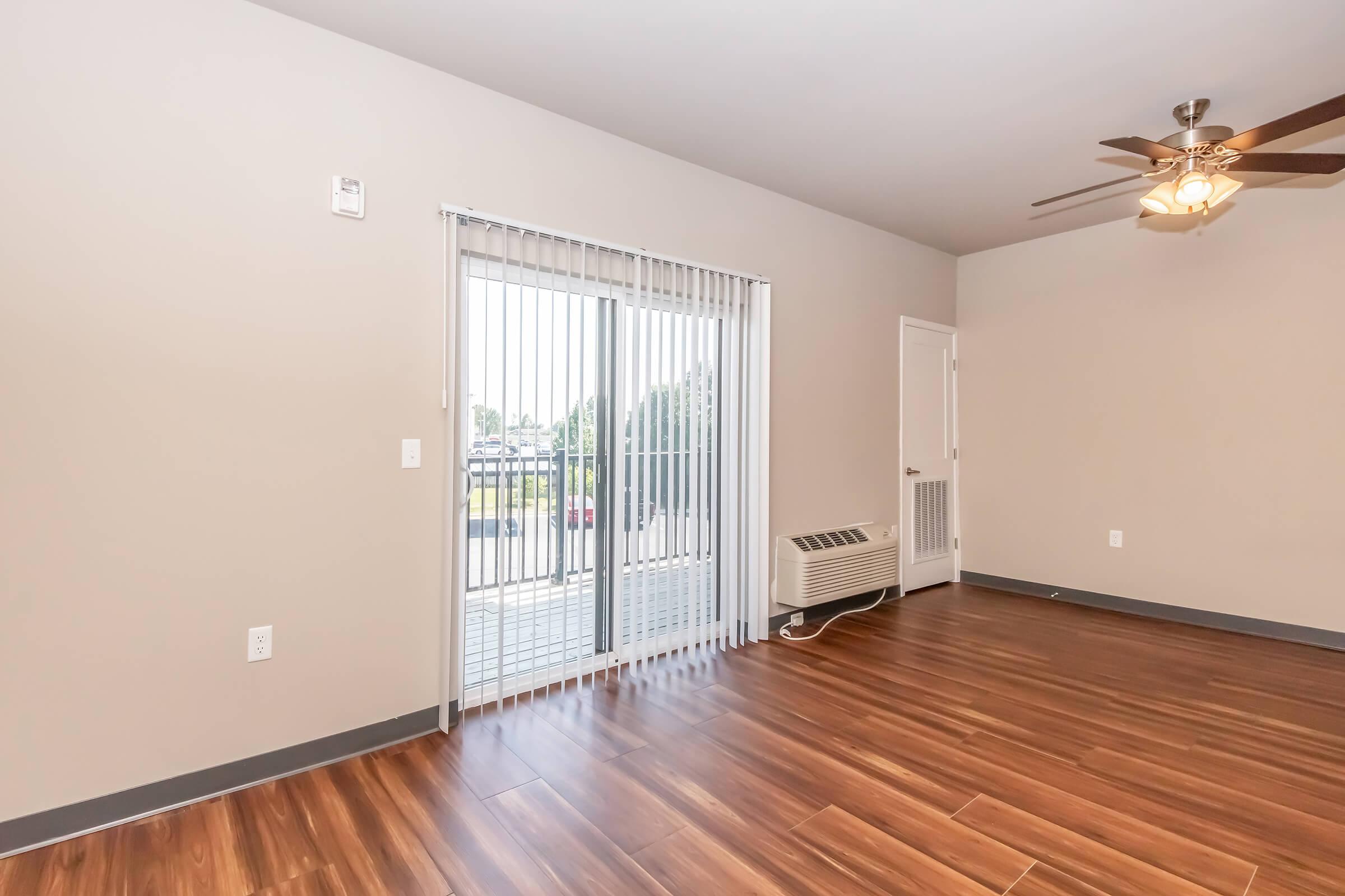 Interior view of a room featuring a wood-style floor, light gray walls, and a ceiling fan. There is a sliding glass door with vertical blinds leading to a balcony, and an air conditioning unit on the floor. The space is empty, creating a bright and open atmosphere.
