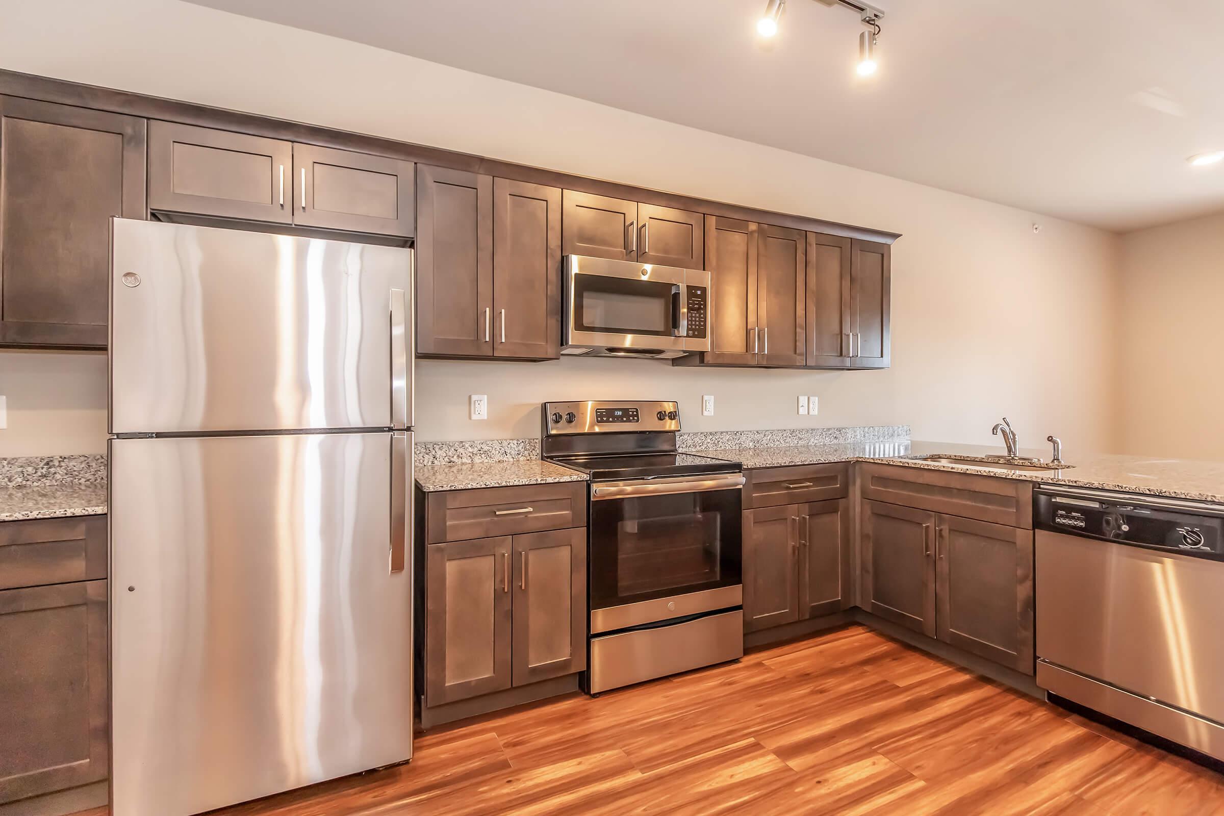 A modern kitchen featuring dark wooden cabinets, stainless steel appliances including a refrigerator, oven, microwave, and dishwasher. The countertops are made of light-colored granite, and the flooring is a warm wood finish. The space is well-lit with overhead lighting.