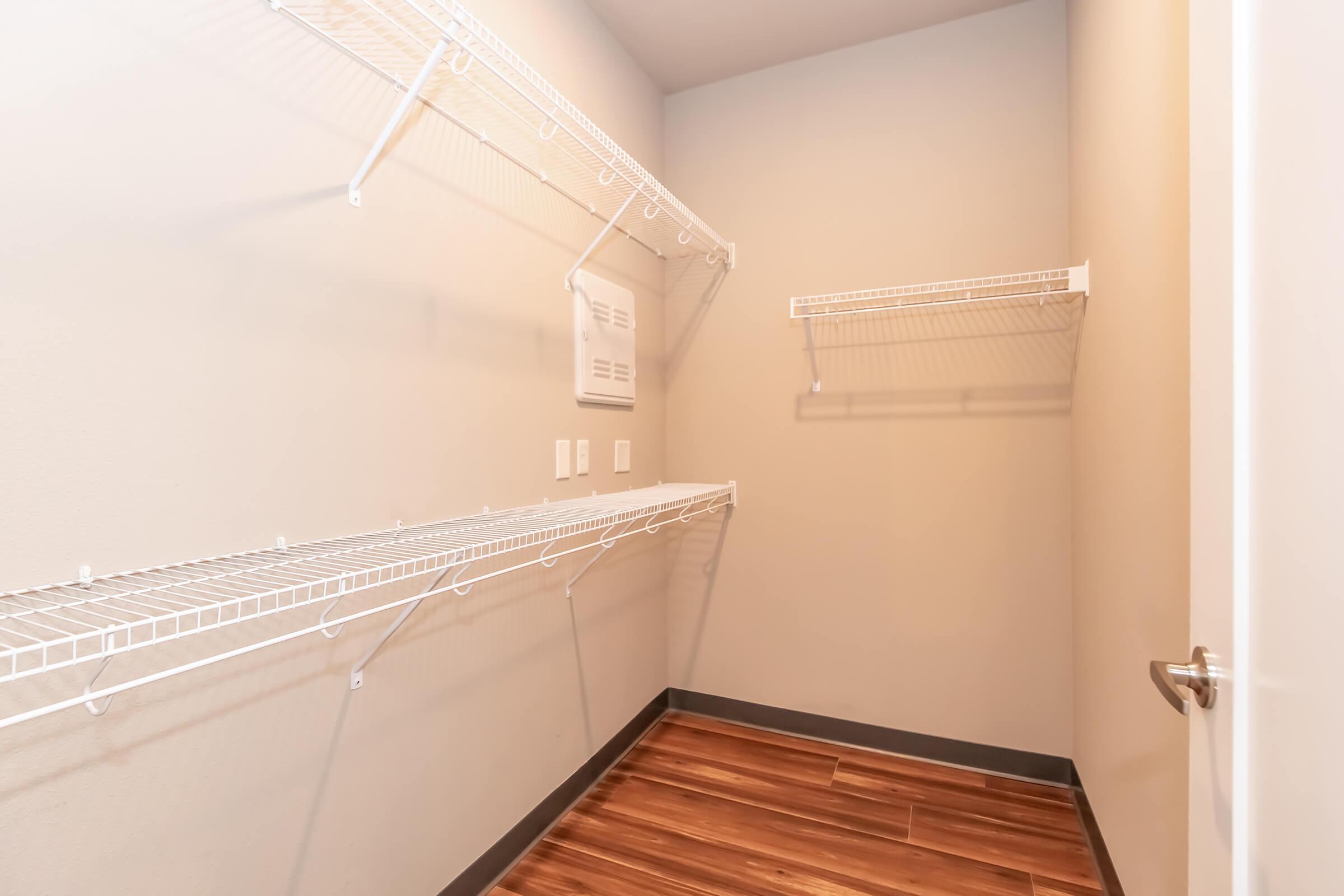 A small, empty closet featuring wire shelves mounted on light gray walls. The floor has dark wooden planks, and there are electrical outlets mounted on one wall. A door is visible on the right side.